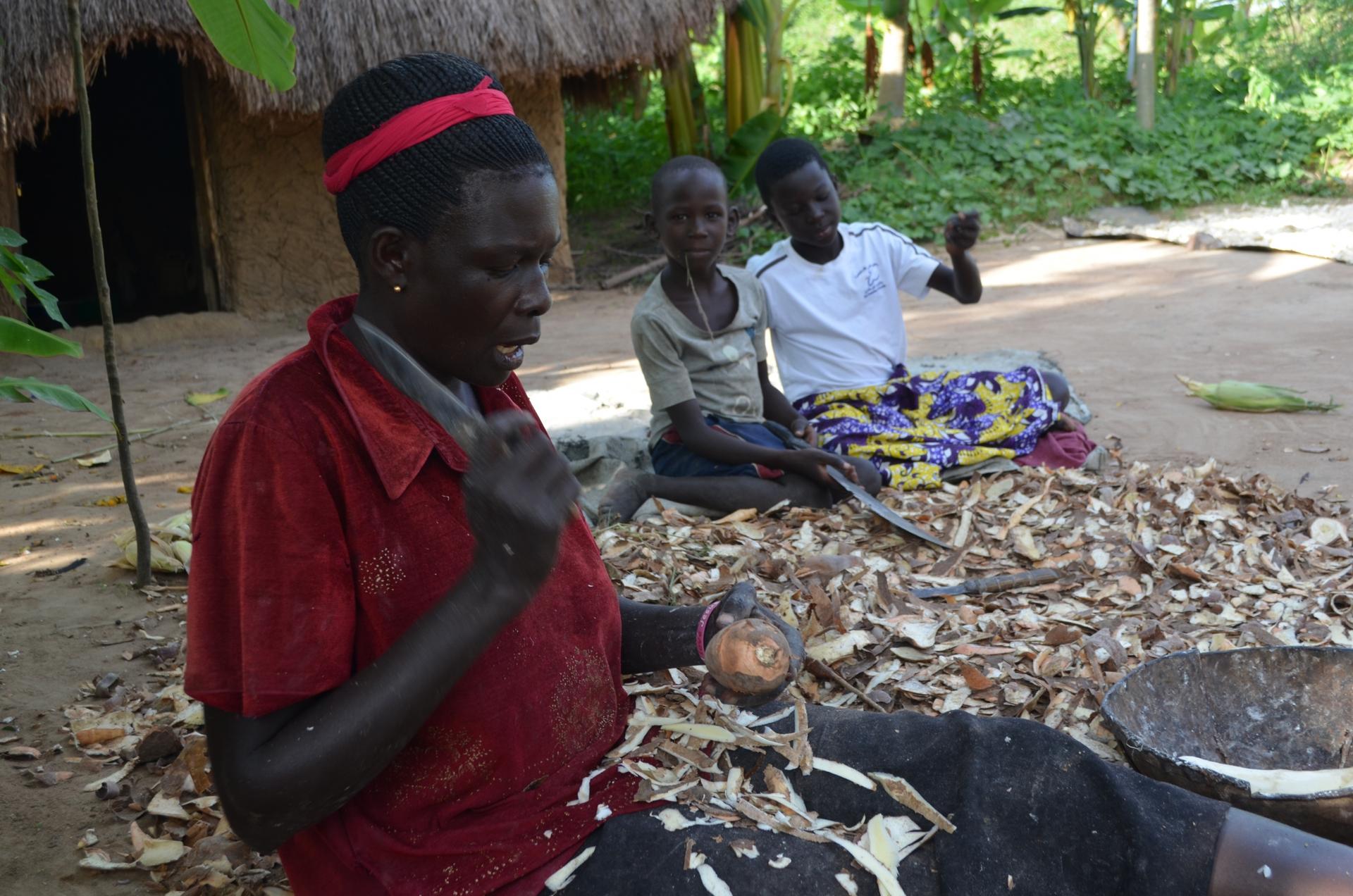 A woman wears a red head band and slices cassava root while sitting on the ground with two children nearby. 
