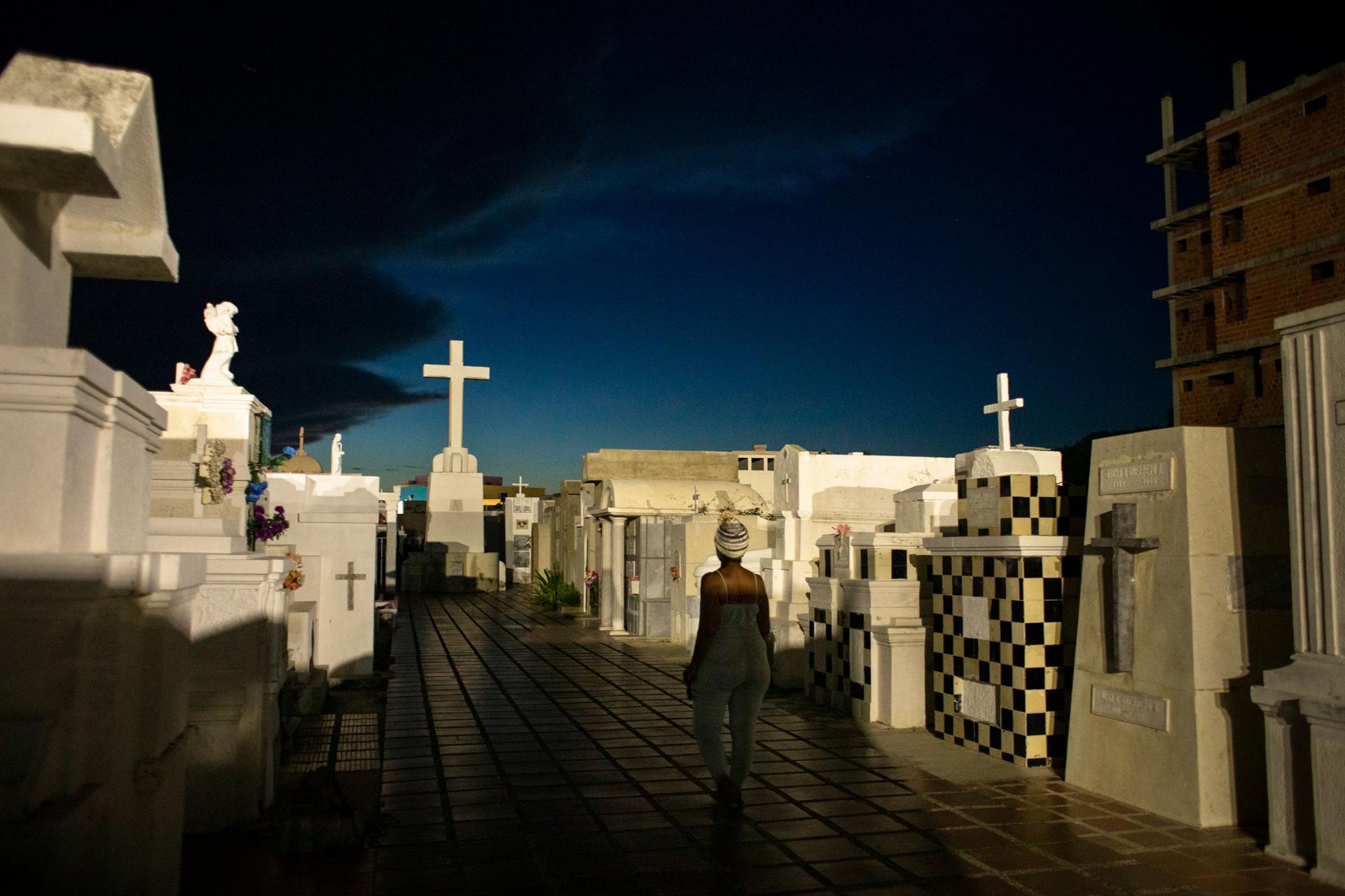 A woman is shown walking in an isle with large tombstones on either side and a white cross at the end.