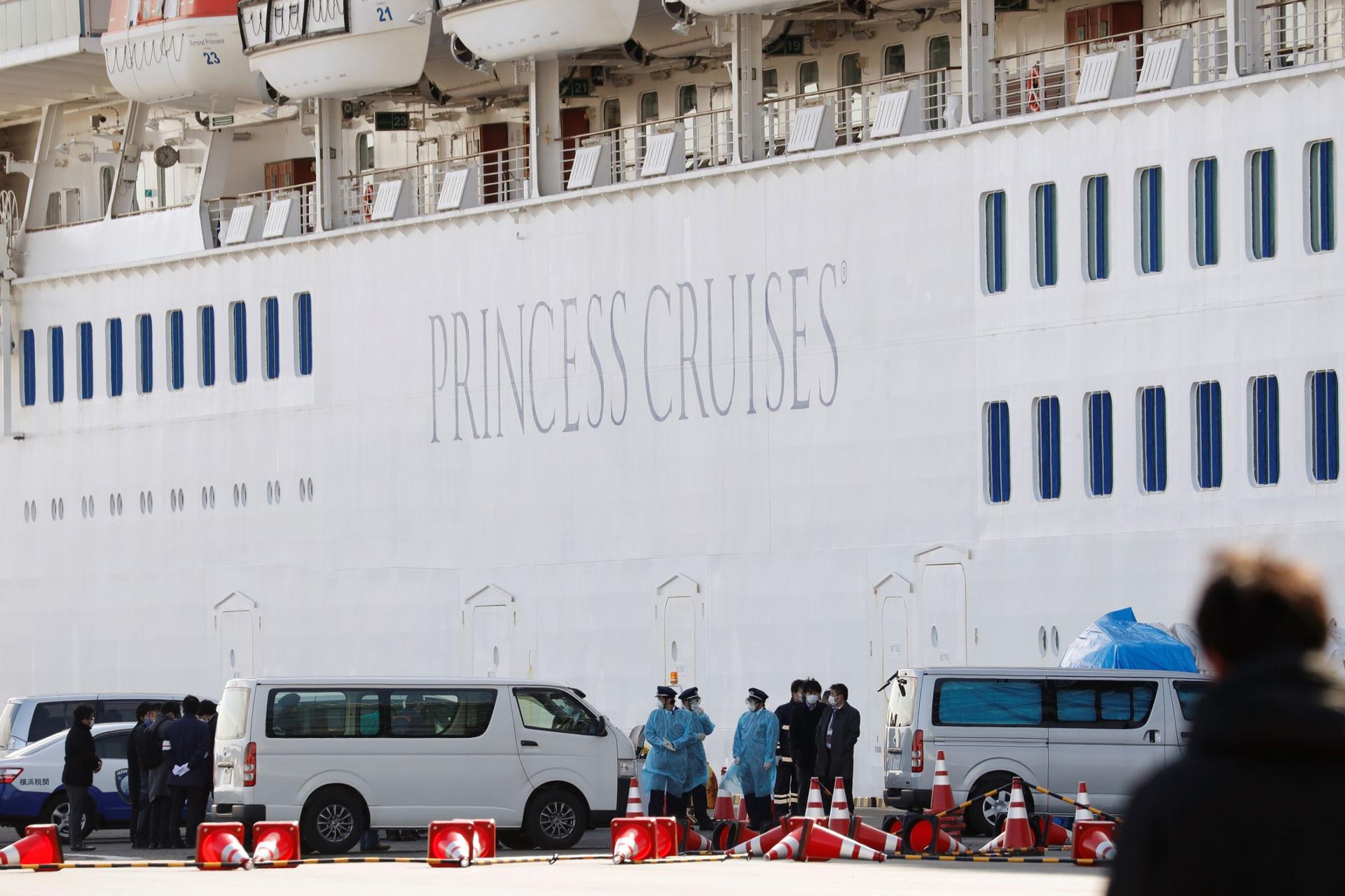 Several vans are shown parked next to a cruise ship with personnel wearing masks stand nearby, 