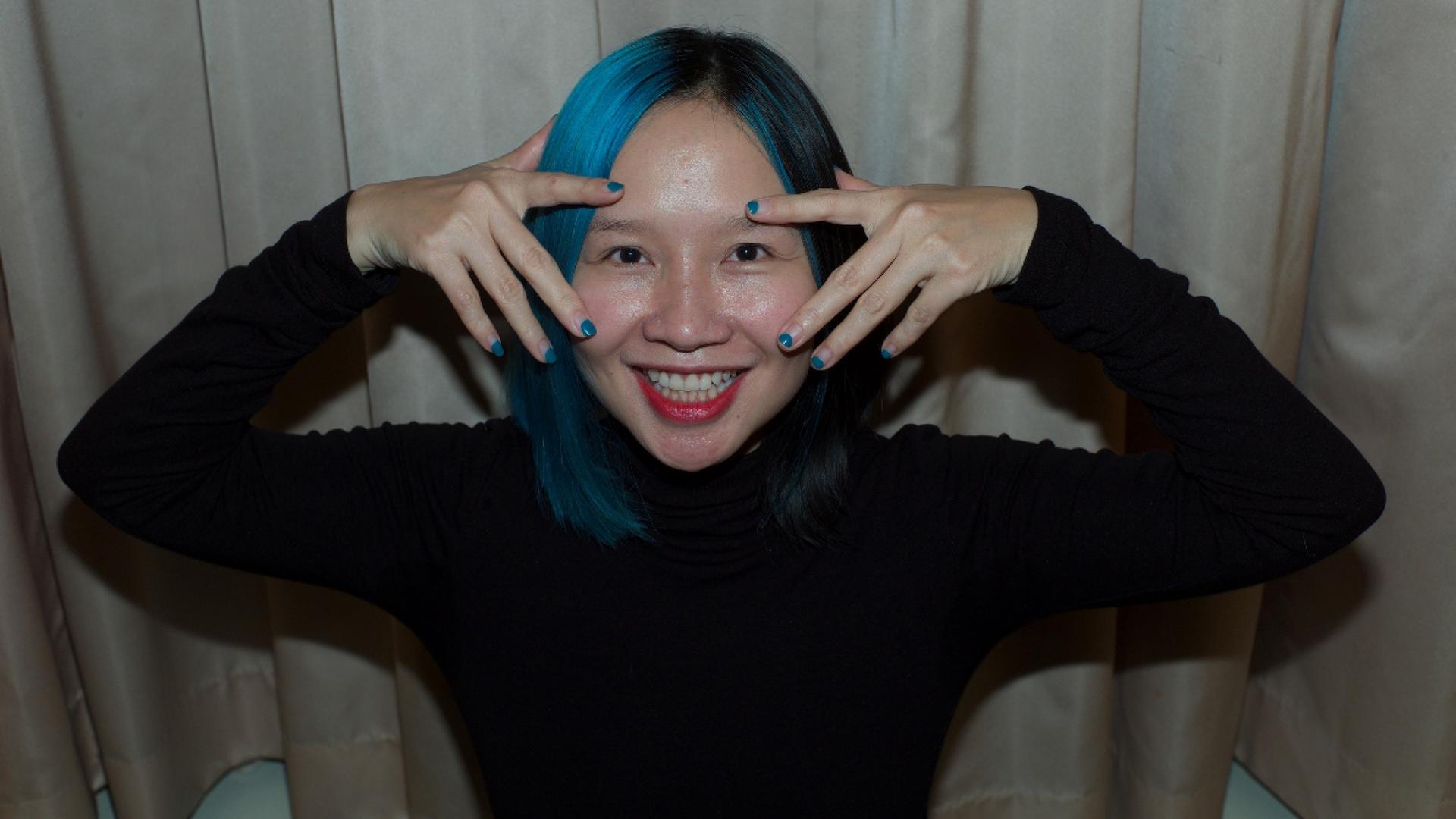 A voman with her hair painted blue holds both or her hands by her eyes.