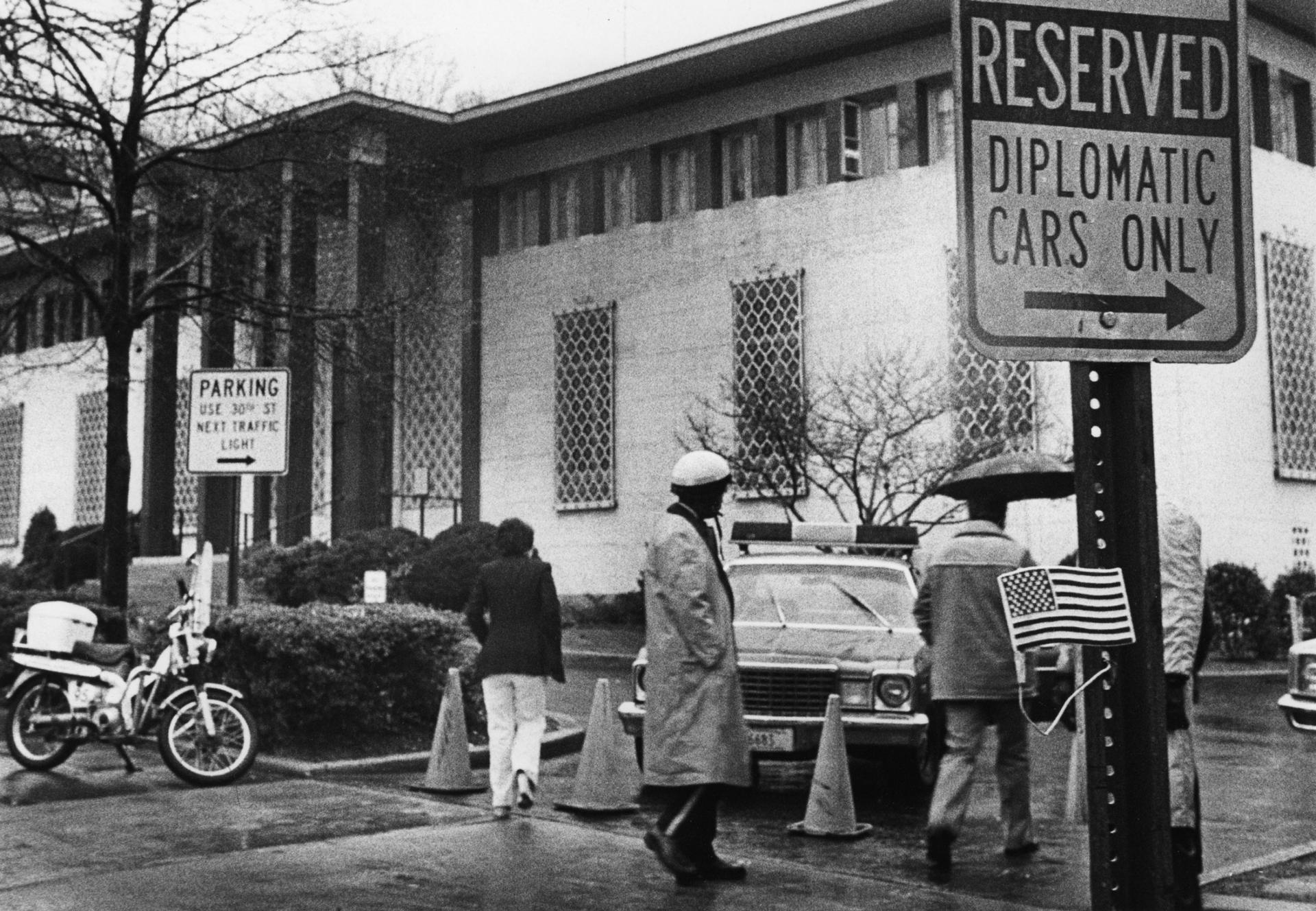 Several people and a police car are shown in this black and white photograph with the former Embassy of Iran in the background.