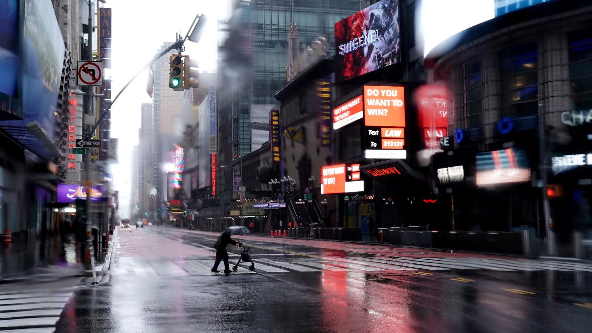A person with a walker crosses and carrying an umbrella is seen crossing 42nd Street with brightly lit signs all around.