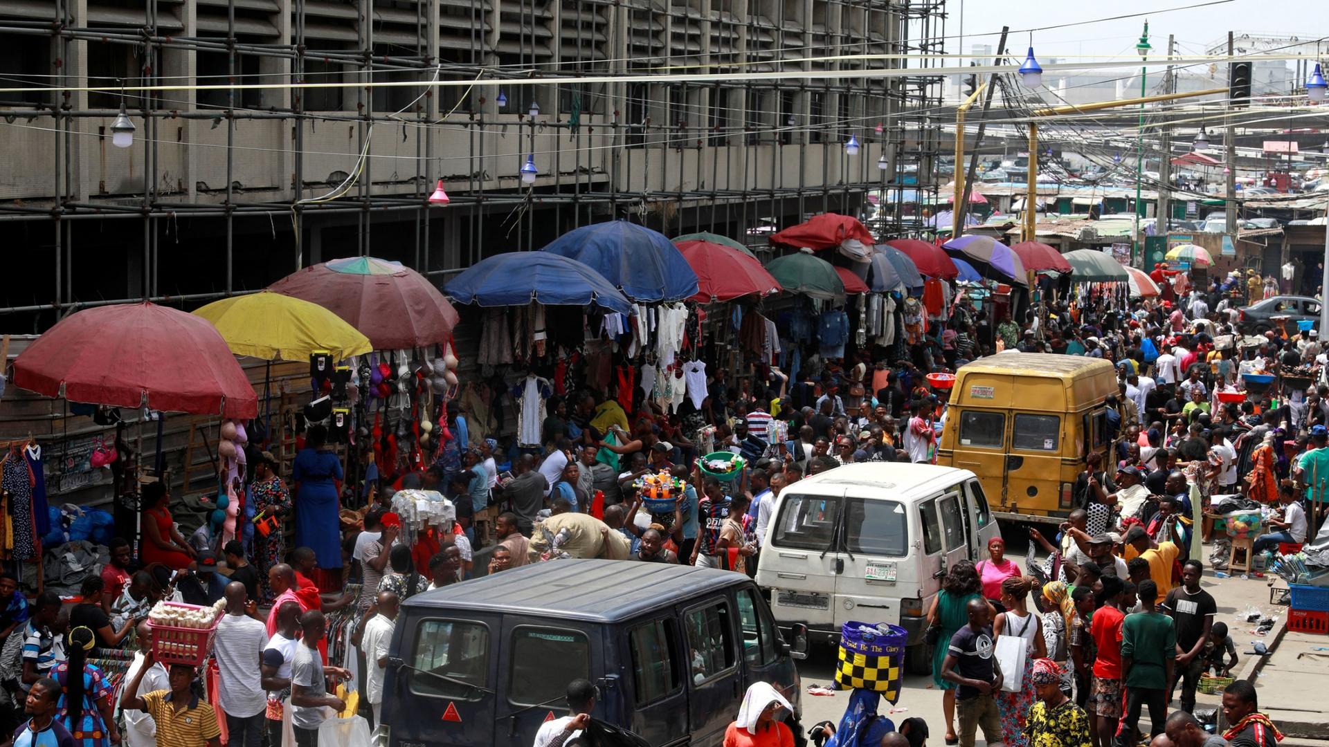A bustling market is shown with hundreds of peope packed into the street lined with large vendor umbrellas.