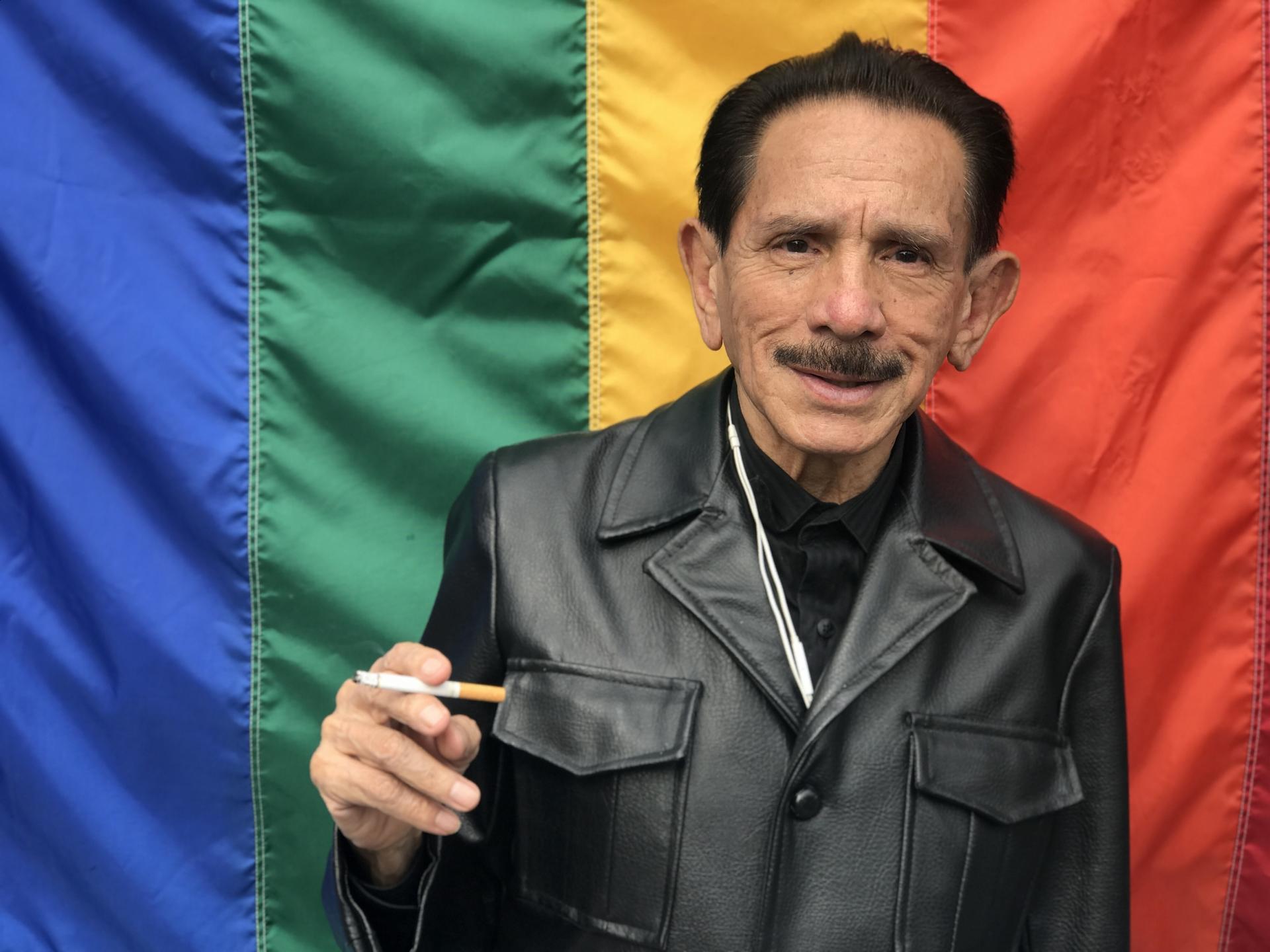 A man wearing a leather vest smokes in front of rainbow flag