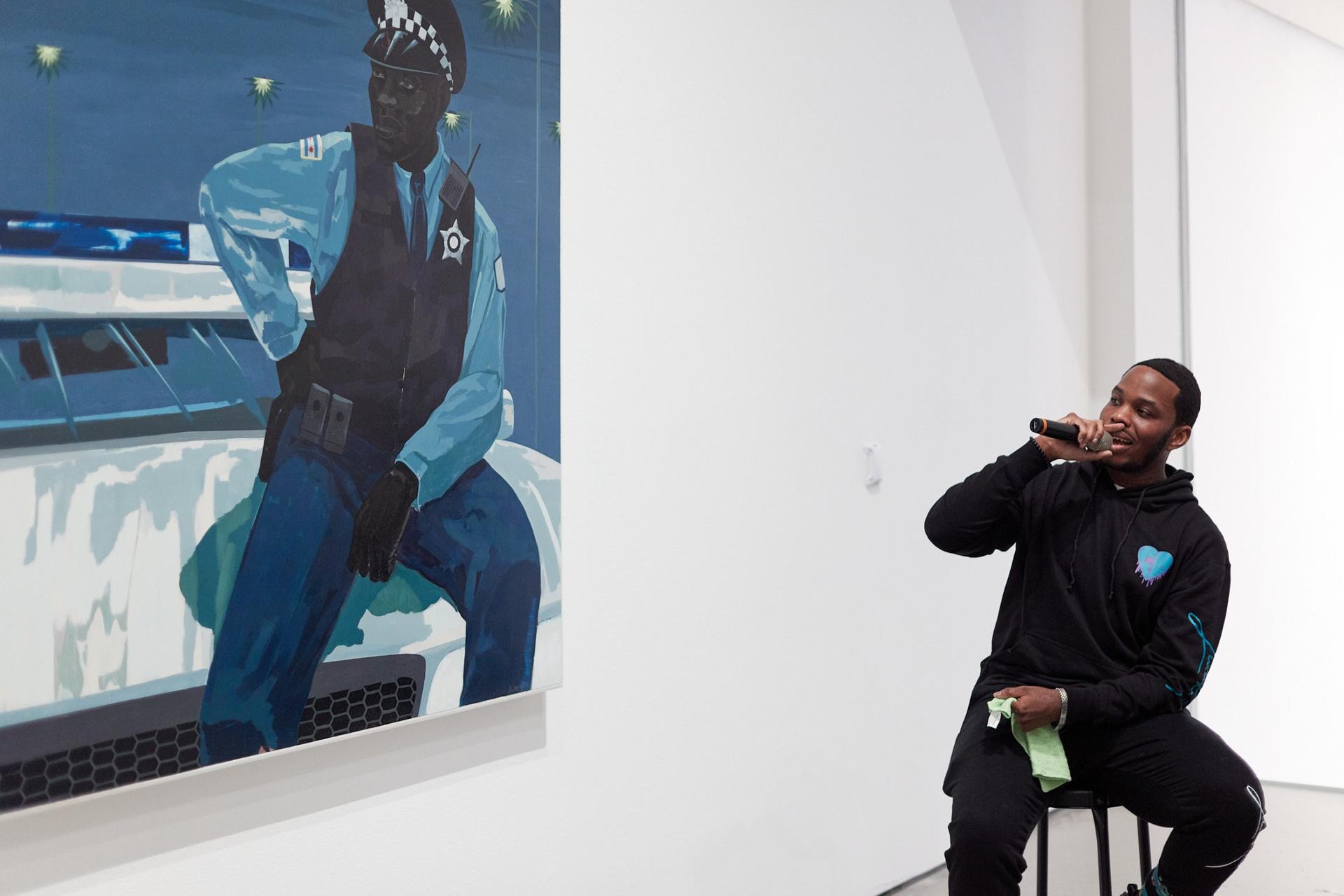 A man in a hoodie sits on a barstool holding a microphone as he looks to a blue-hued painting of a policeman. 