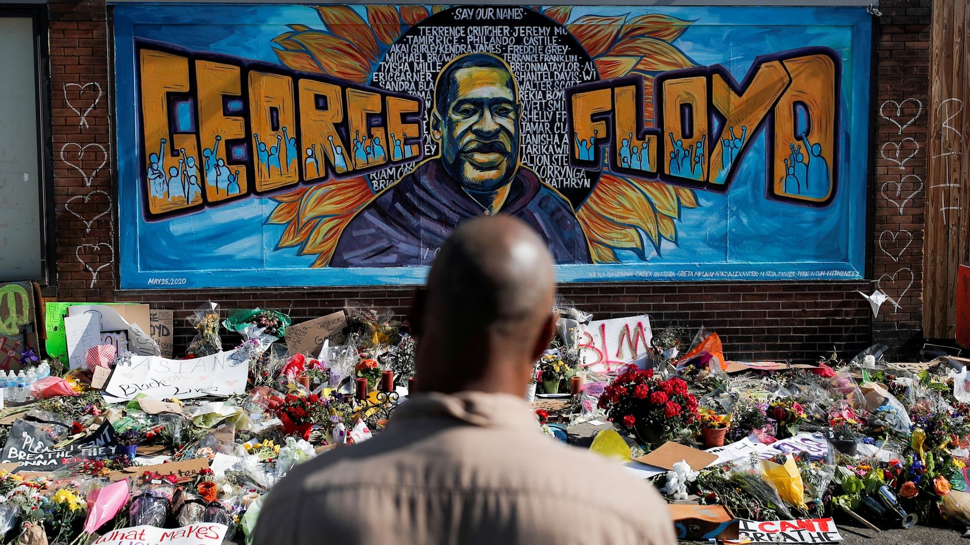 A black man stands in front of a mural honoring George Floyd and other black Americans killed by law enforcement