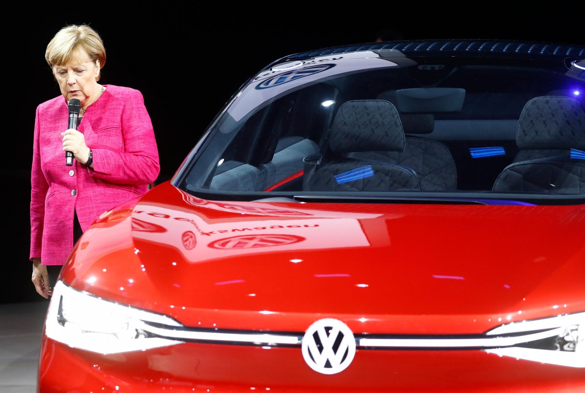 German chancellor Angela Merkel is shown wearing a pink jacket and standing next to a red VW car.