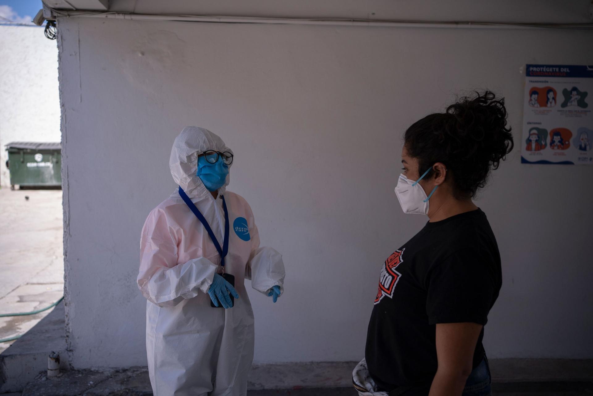 A woman is shown wearing fully covering protective medical clothing and standing across from another woman wearing a white face mask and dark t-shirt.