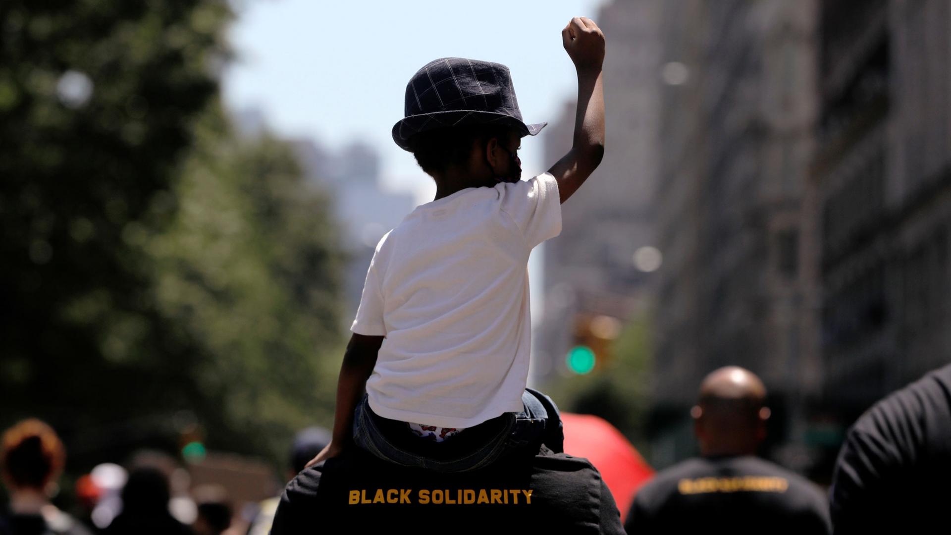 A young boy is shown wearing a white t-shirt and had while sitting on the shoulder of a man wearing a shirt with "Black Solidarity" printed on it.