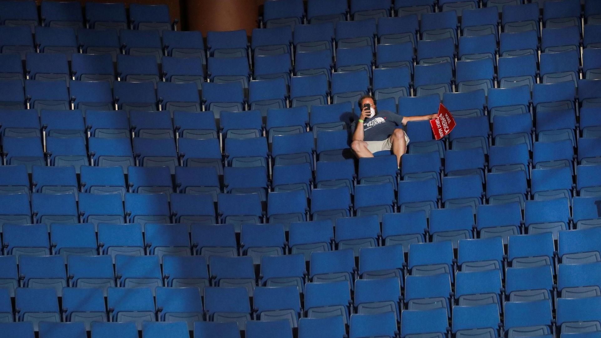 A man is shown sitting in a chairi in an arena hollding his phone up with empty rows of chairs all around him.