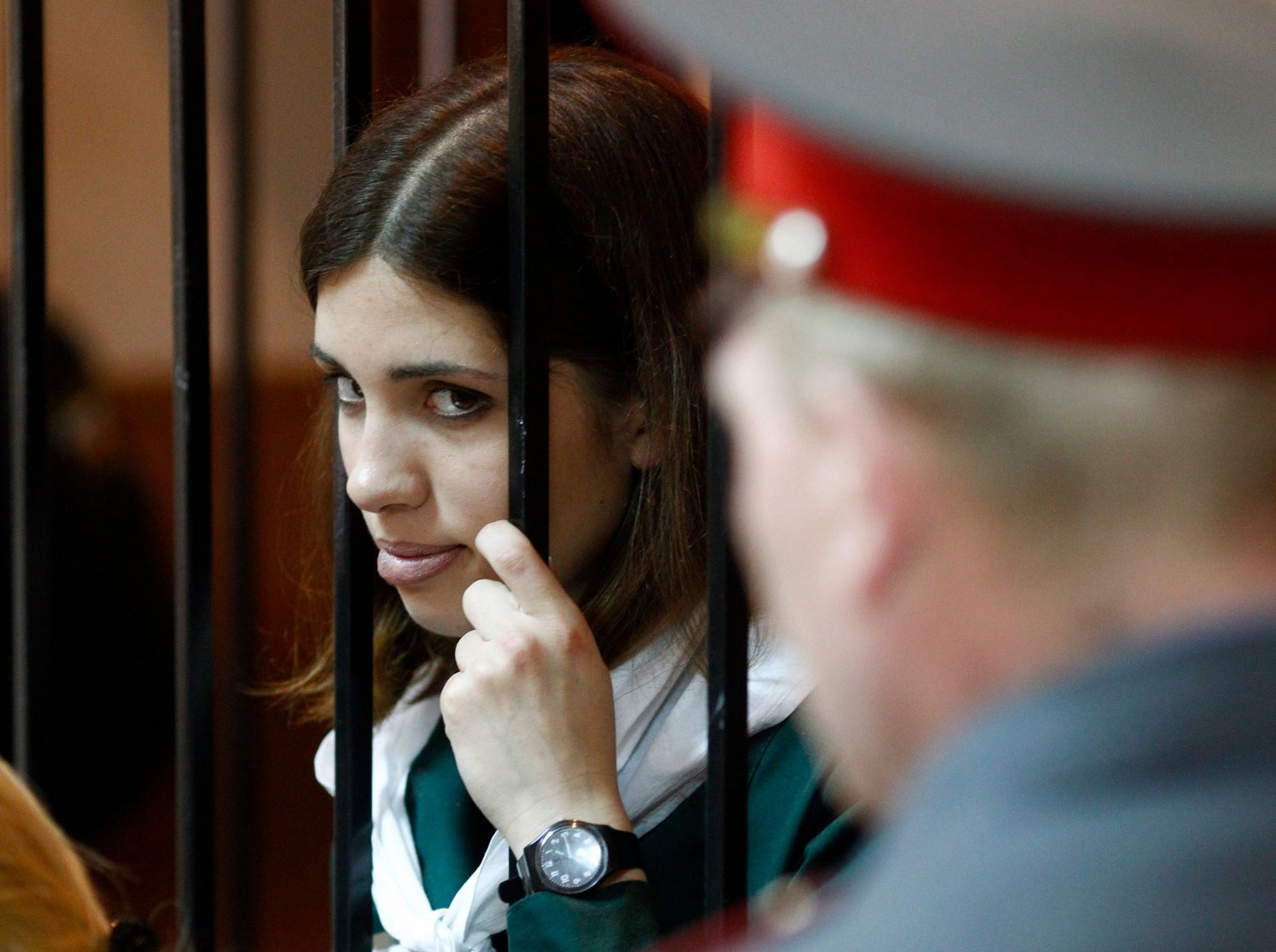 A young woman holds bars of a cell and peers out in a courtroom 