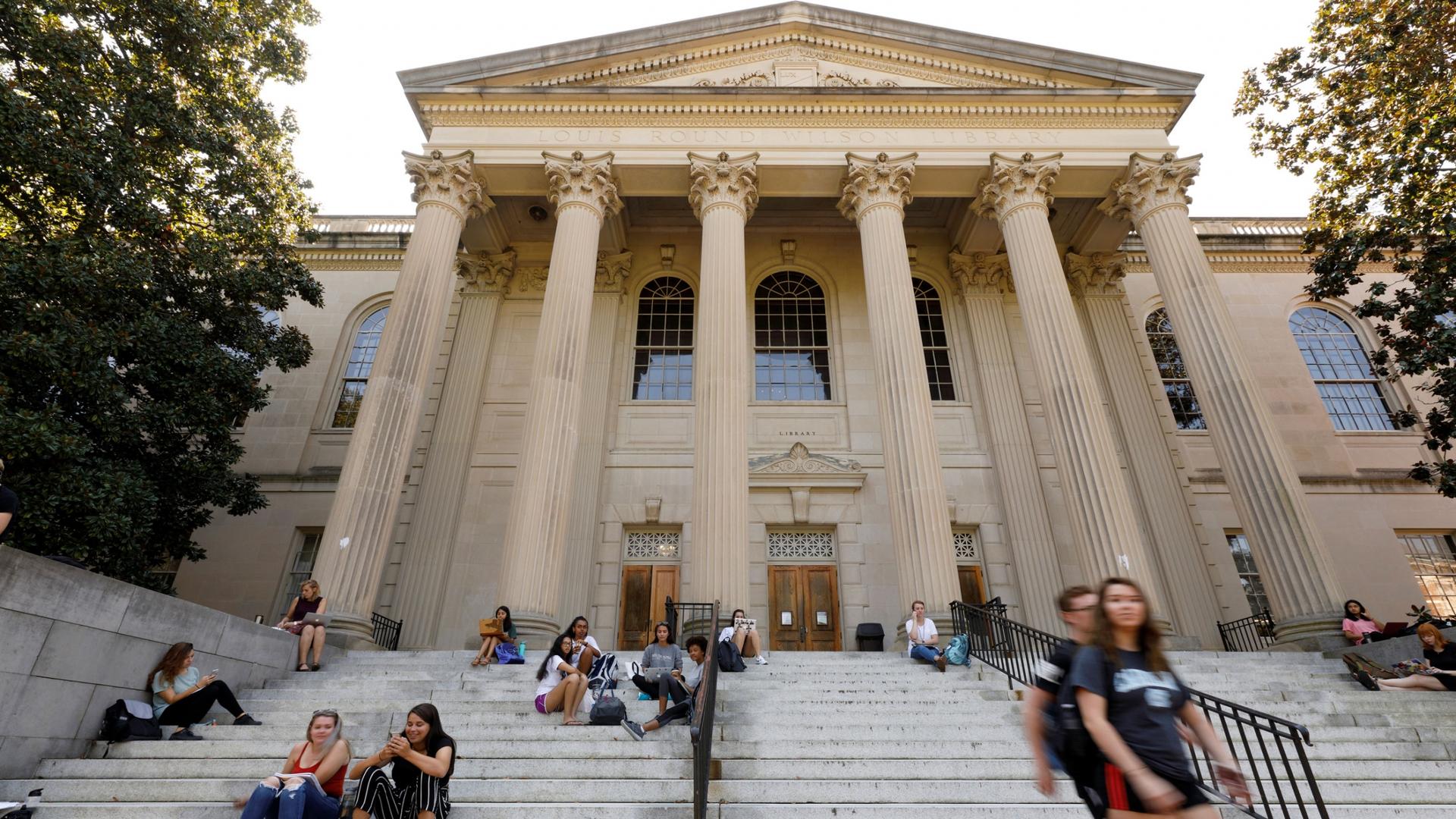 Several students are show sitting on steps with the large Greek columns at the entrance of a college library are shown in the distance.