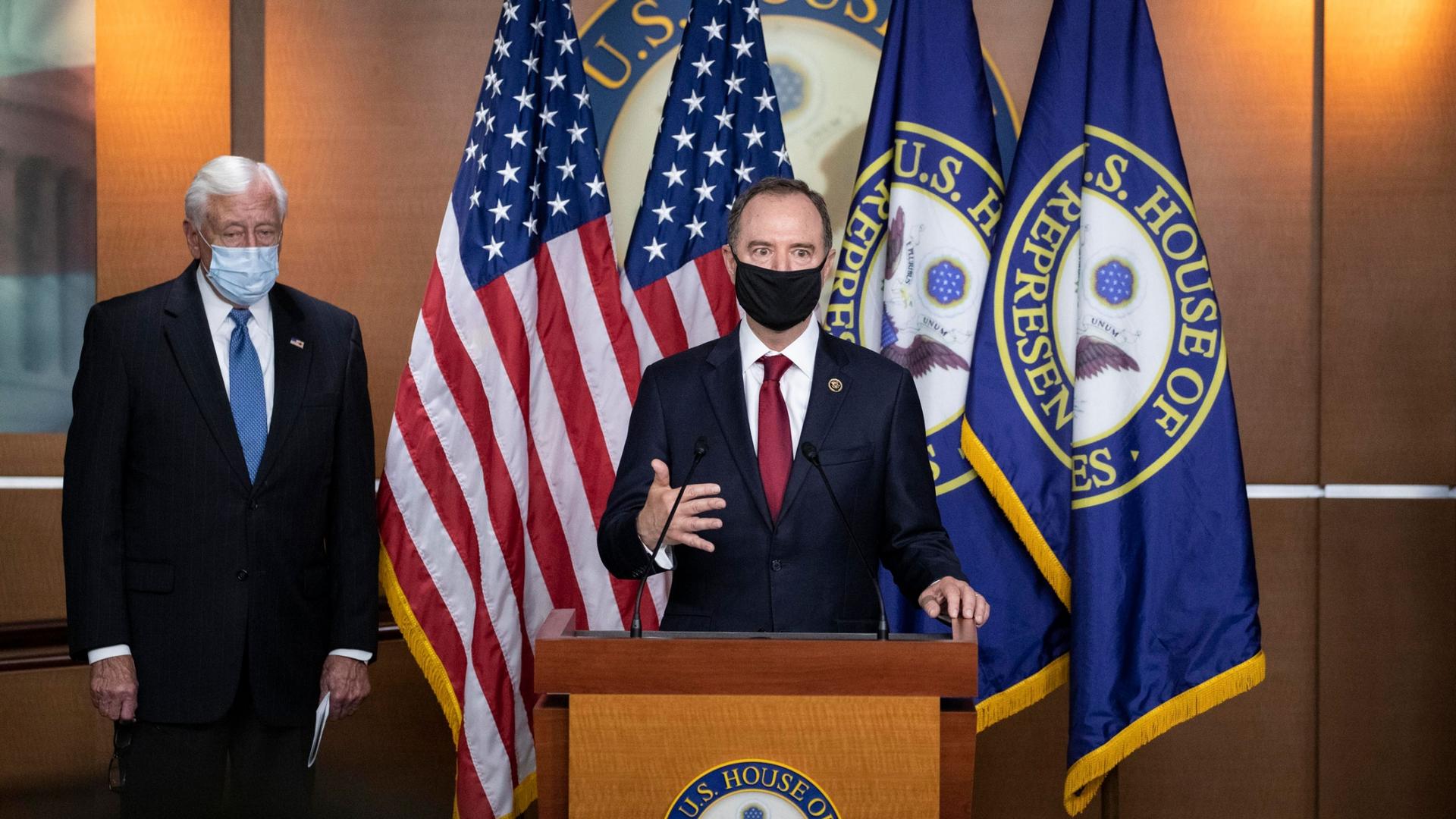 Rep. Adam Schiff is shown wearing a black face mask and standing at a wooden podium with flags behind him.