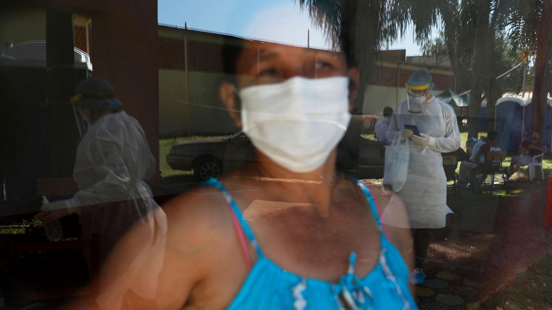 A woman is shown with a face mask and wearing a blue top and looking through glass where several people are shown wearing full medical protective gowns.