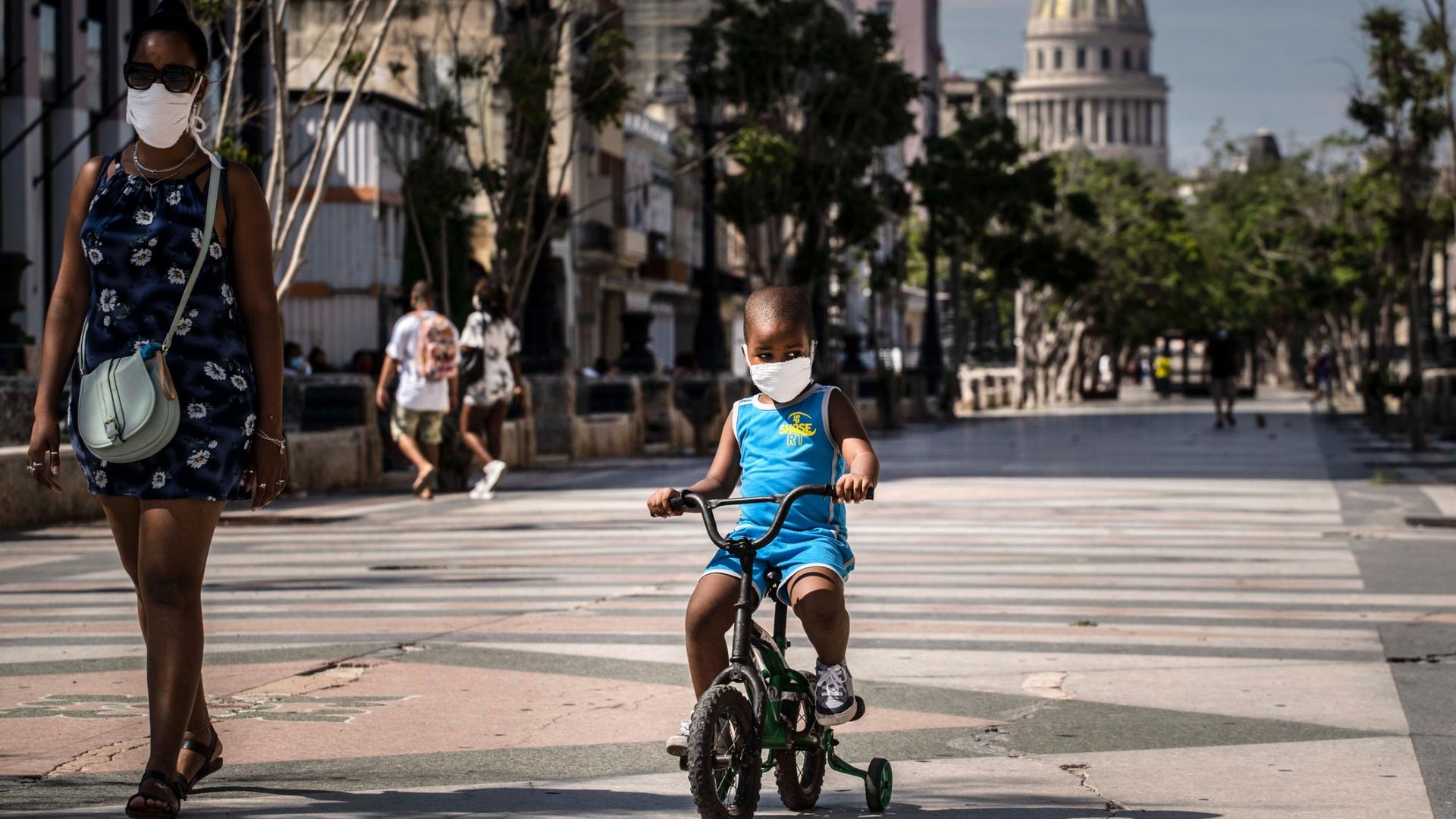 A young child is shown wearing a face mask and a blue outfit and riding a small bicycle with training wheels.