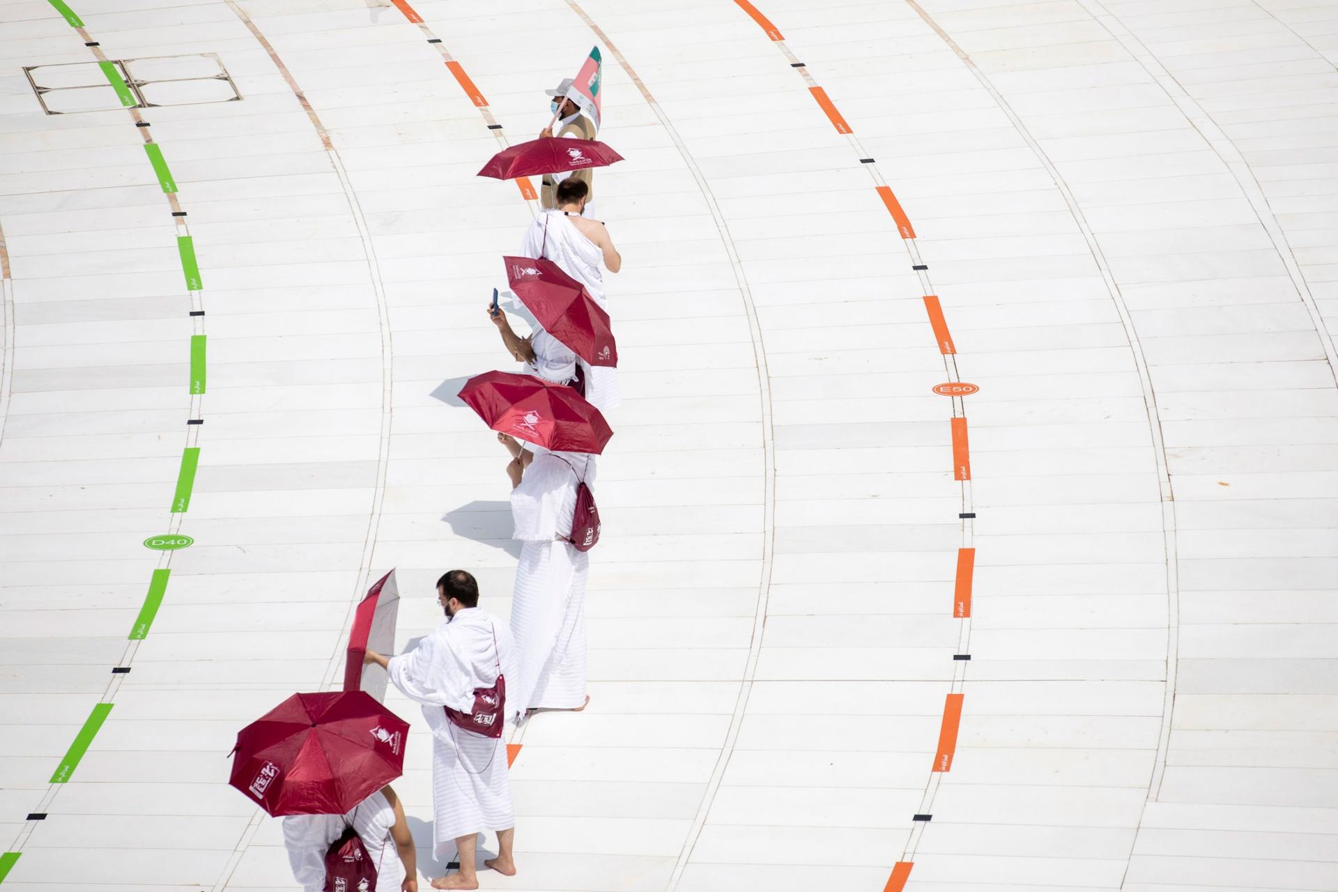 A line of people are shown wearing white robes and carrying red umbrellas.