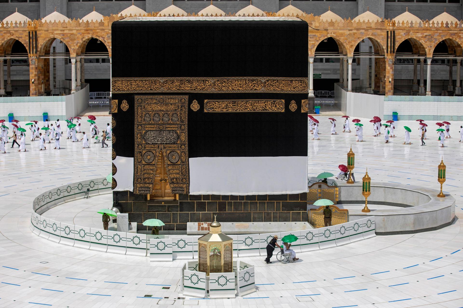 The cube-like Kaaba is shown center frame with a pilgrim being pushed in a wheelchair in the nearground.