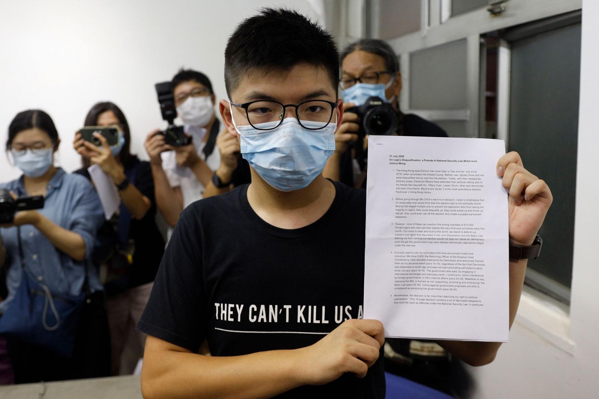 Hong Kong pro-democracy activist Joshua Wong is shown wearing a face mask a glasses while holding a white document up to the camera.