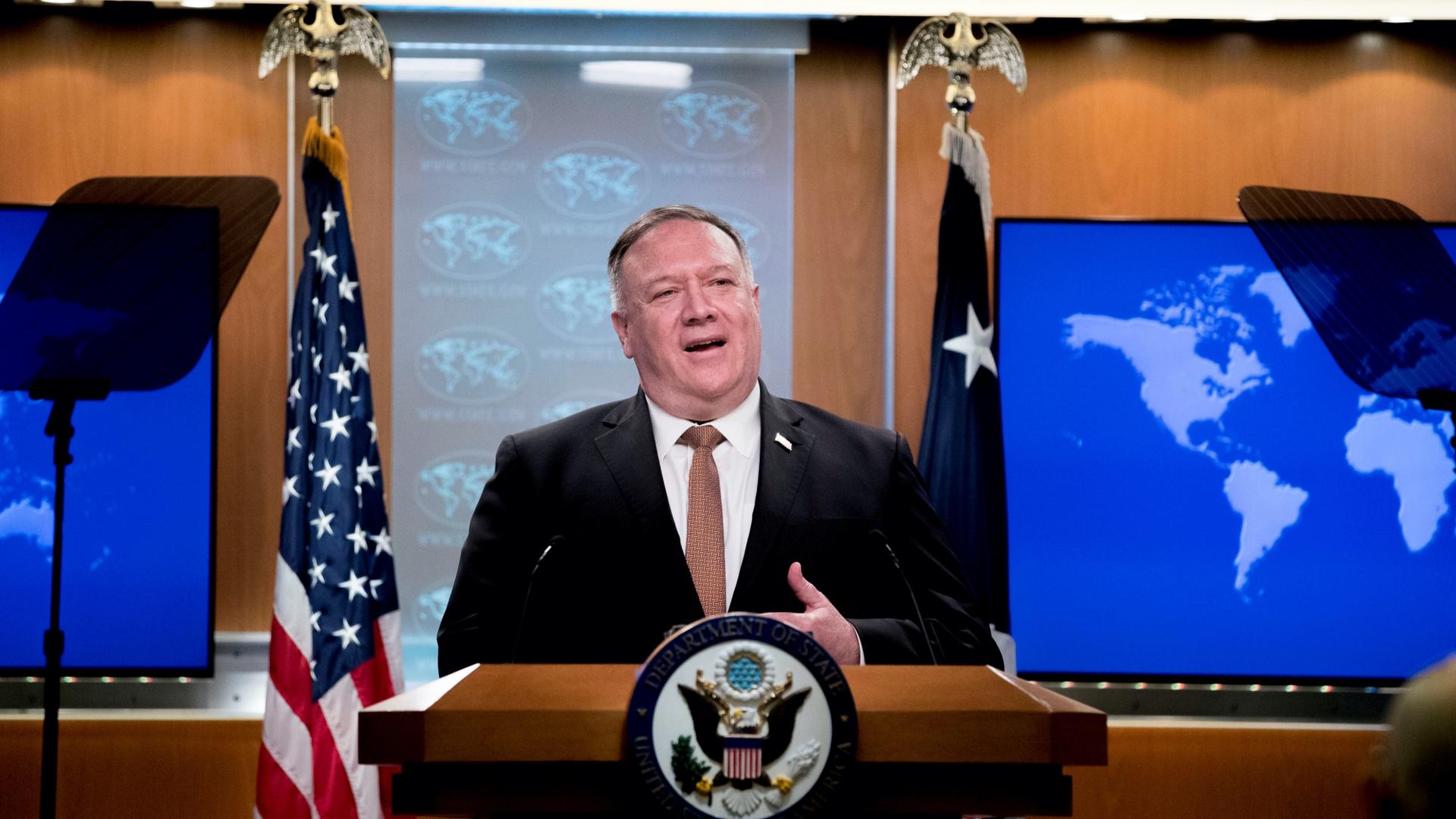 A man wearing a suit stands at a State Department podium between two flags 