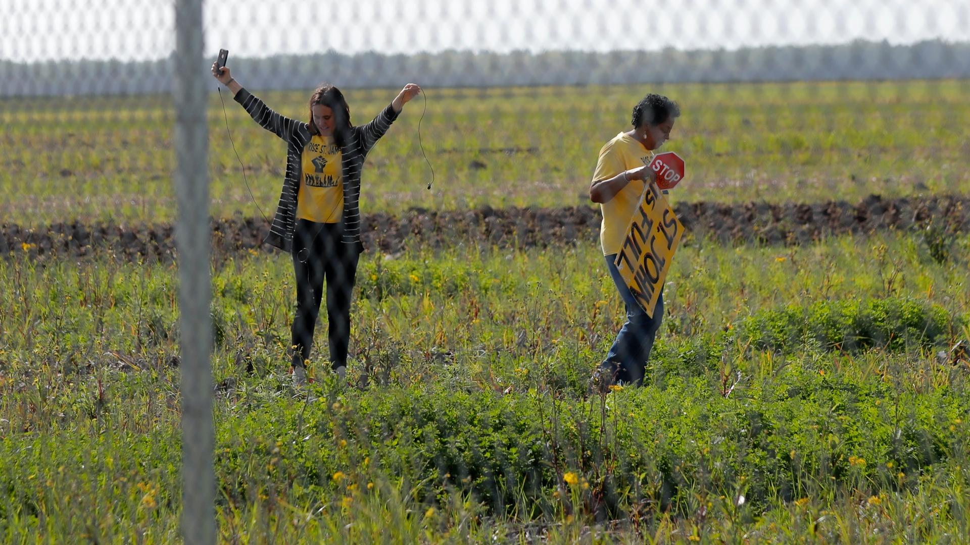 Two Black American women wearing yellow activist shirts stand in a field behind a fence 