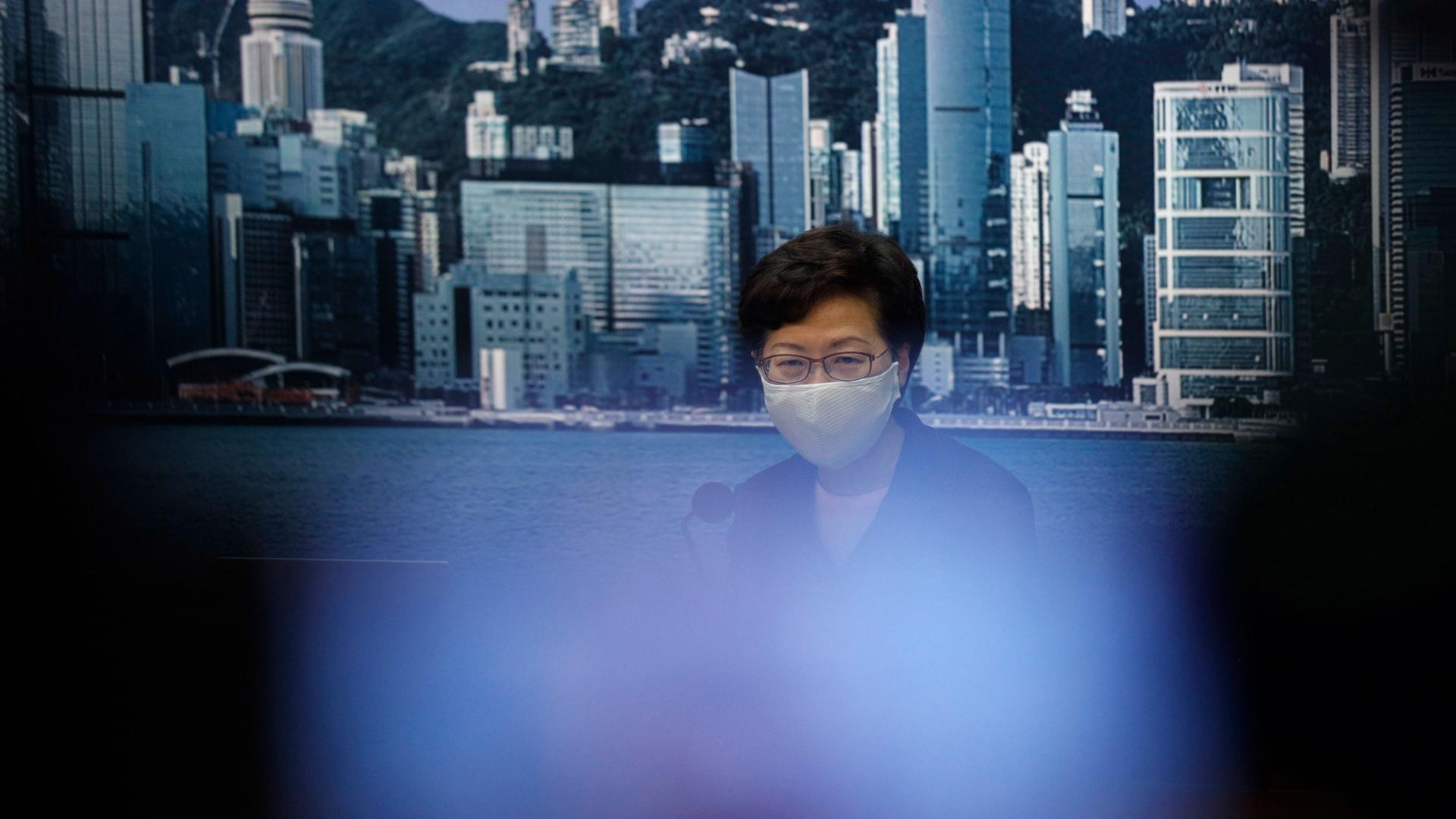 Hong Kong Chief Executive Carrie Lam is shown wearing a face mask with an image of the Hong Kong coastline in the background.