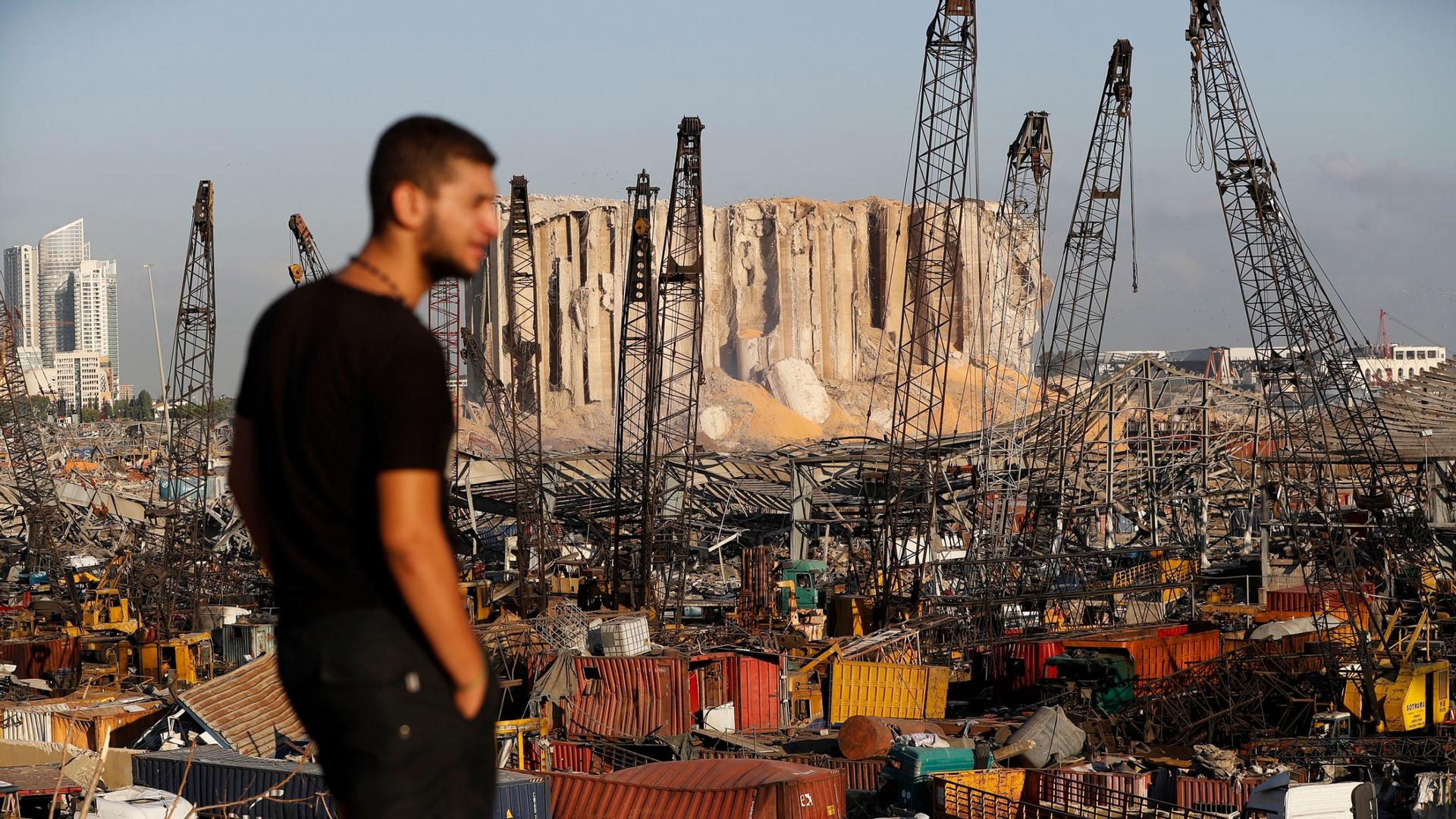 The port of Beirut is show with shipping containers scattered and damaged in the distance with a man in a dark t-shirt in the nearground.