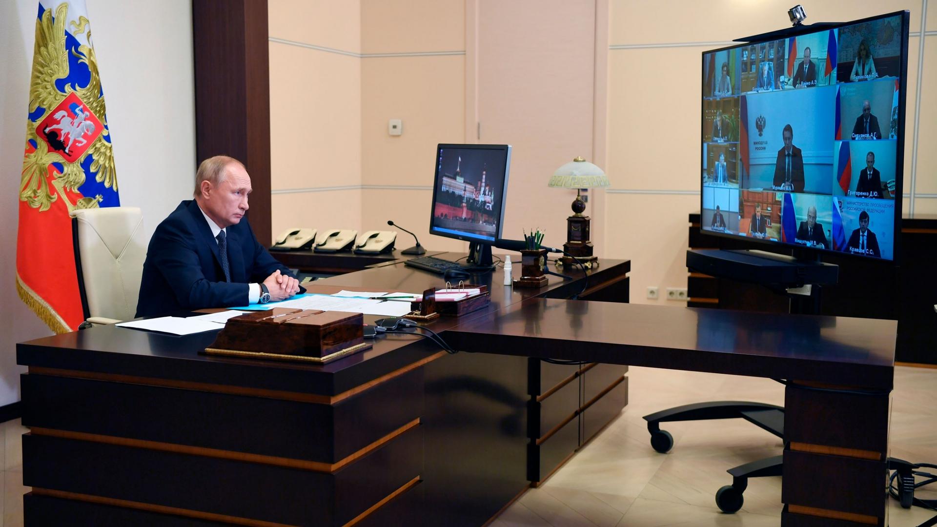 Russian President Vladimir Putin attends a virtual cabinet meeting while sitting at a large wooden desk with a television monitor showing several officials.