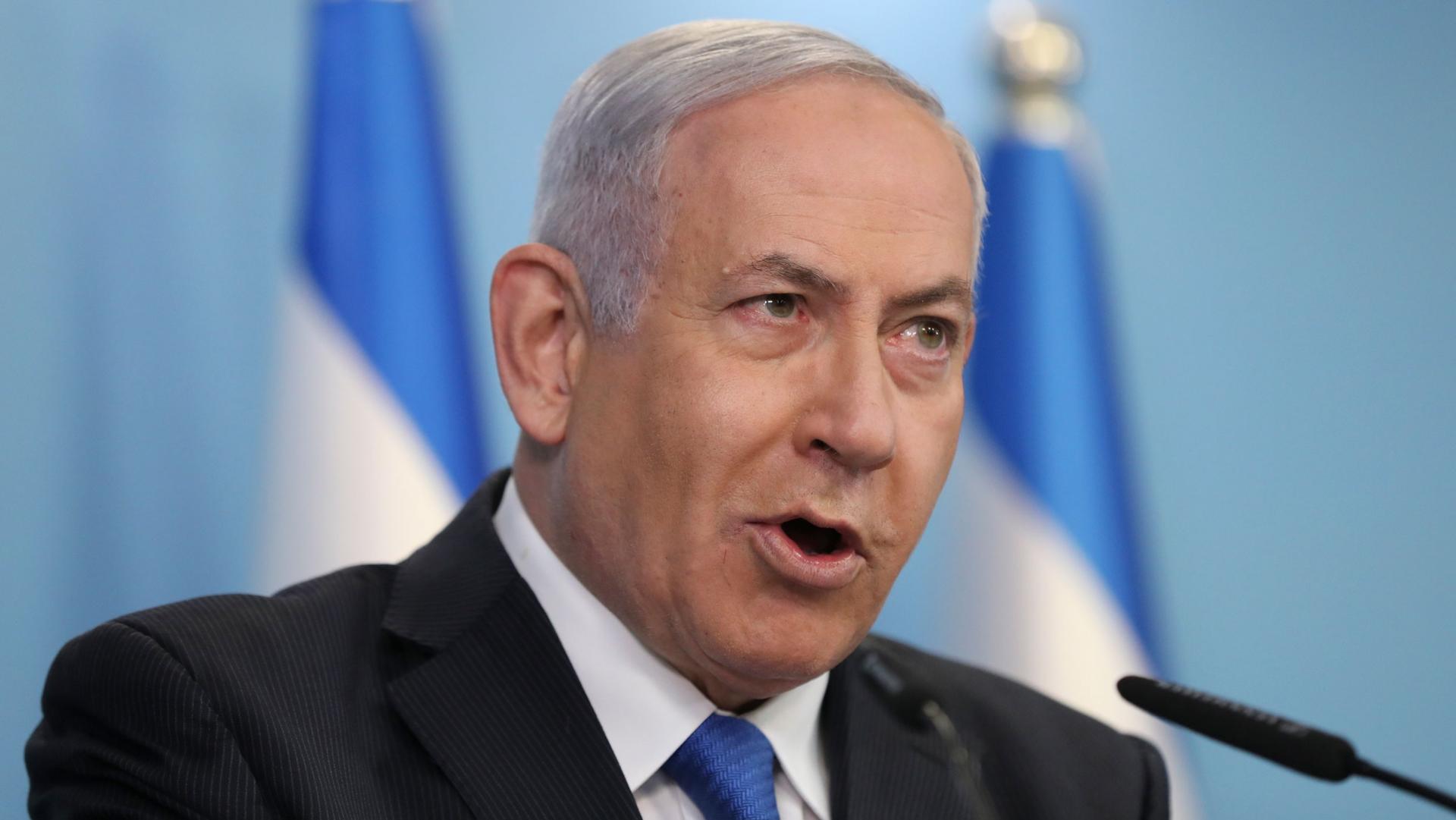 Israeli Prime Minister Benjamin Netanyahu is shown wearing a dark suit and blue tied while speaking into a microphone.