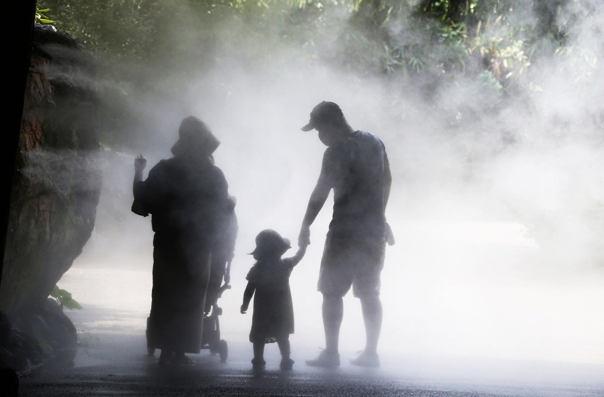 Two people hold the hand of a small child as they are surrounded by mist to cool off.