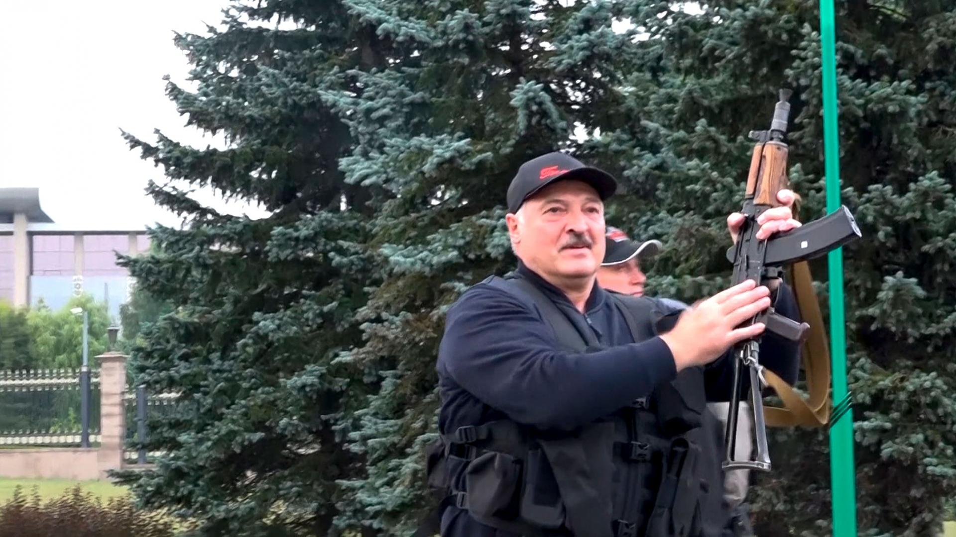 Alexander Lukashenko is shown wearing a hat and black vest while holding an assault rifle in the air.
