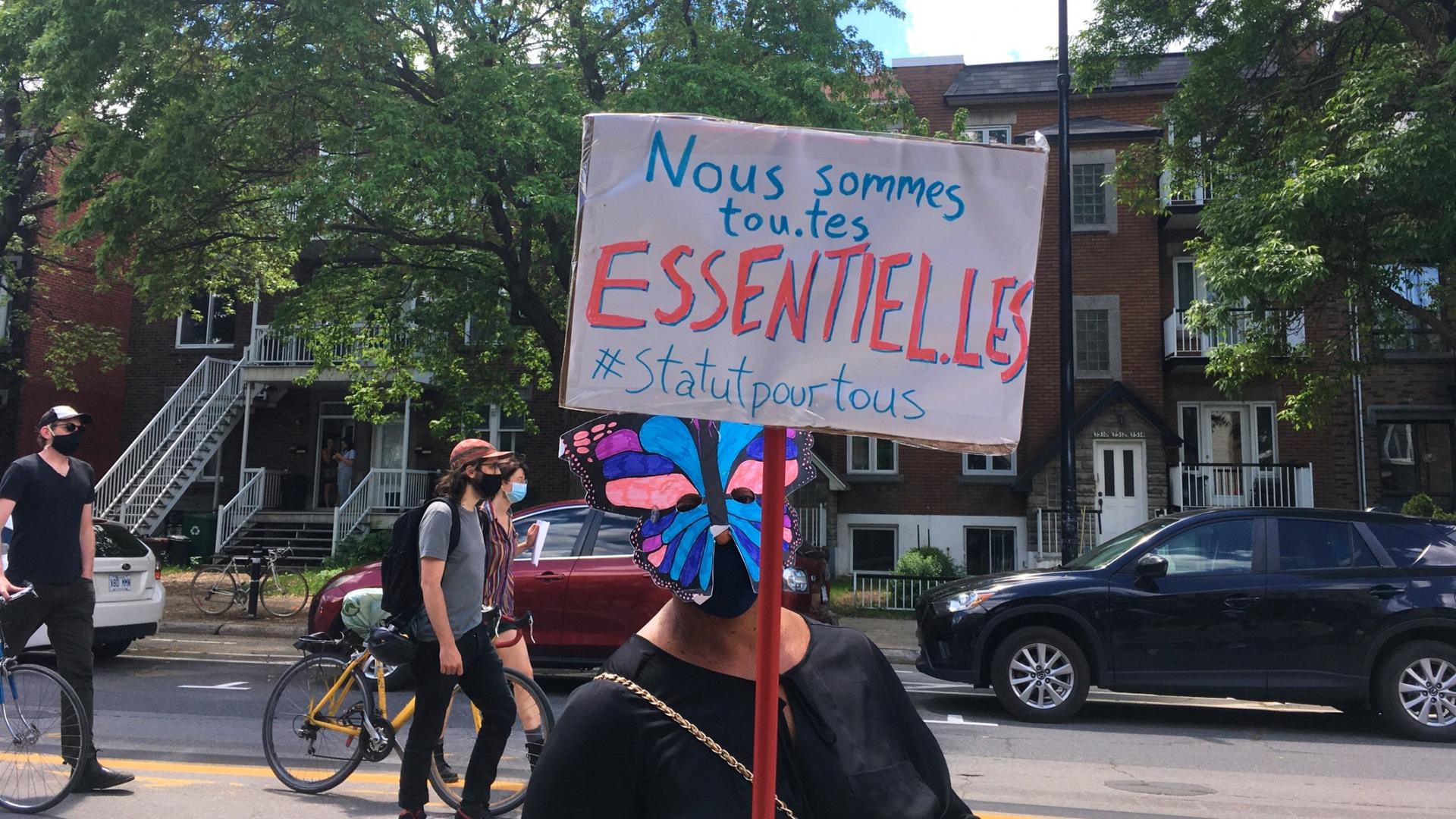 A demonstrator in Montreal holds up a sign in French saying, "We are all essential, status for all."
