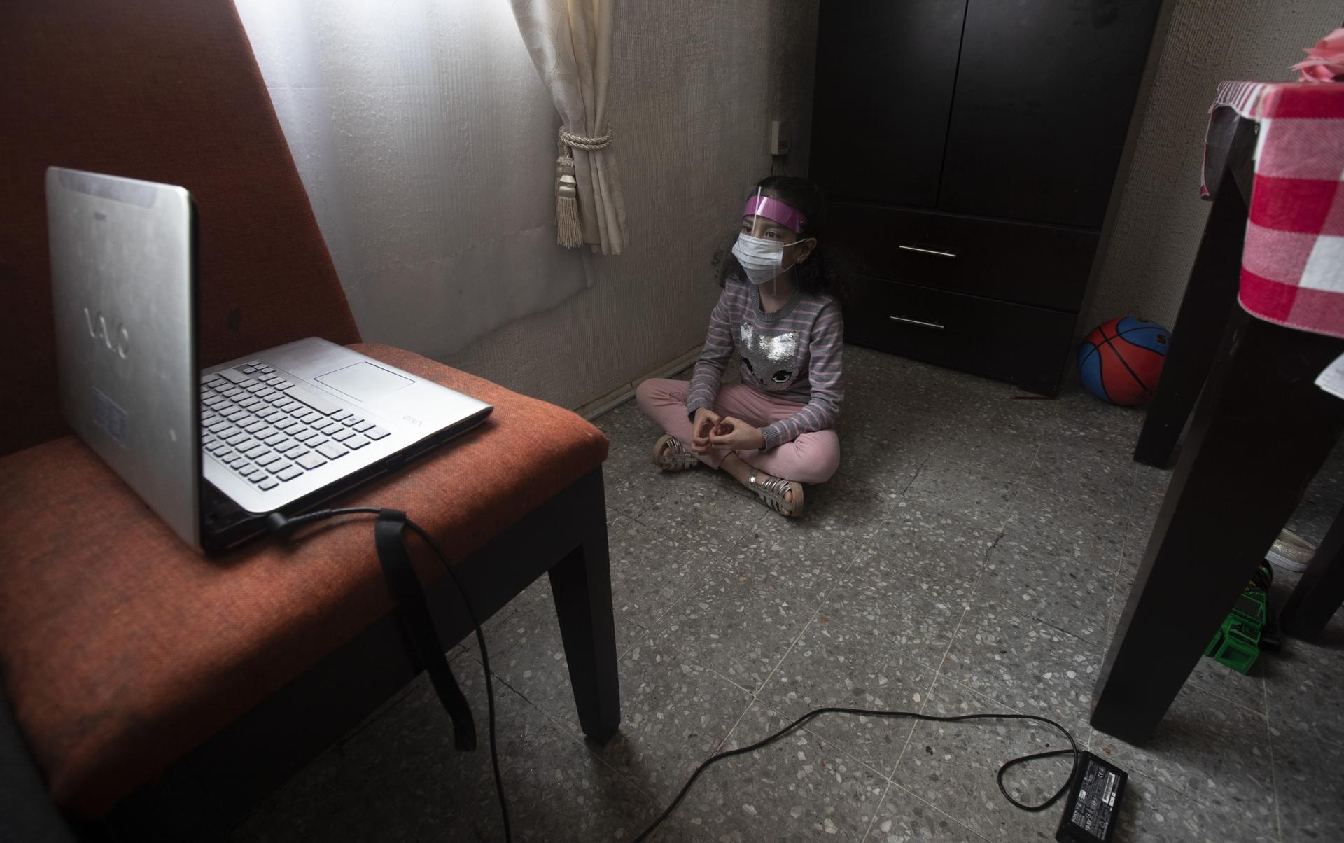 A girl wears a plastic guard indoors as she watches a laptop screen. 