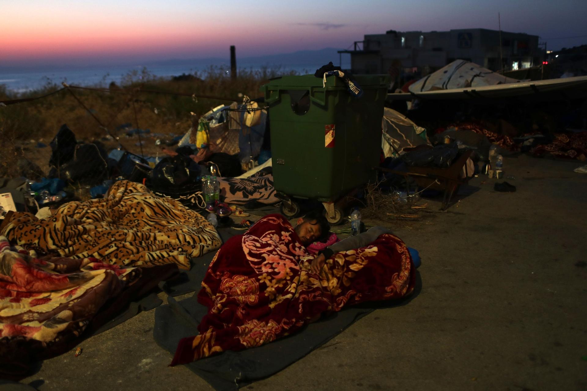 Several migrants and refugees are shown sleeping under blankets on the side of a road with a green trash bin with wheels nearby. 