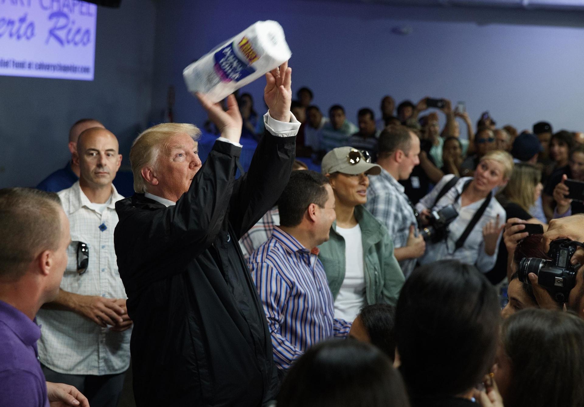 A man wearing a black suit throws a roll of paper towels to people in front of him to catch.