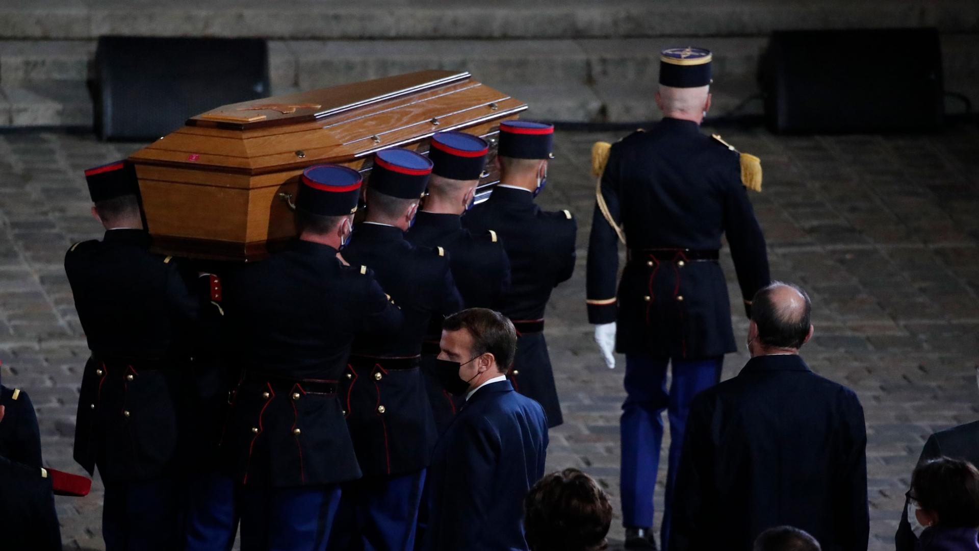 Eight uniformed officers are shown carrying a brown wooded coffin.