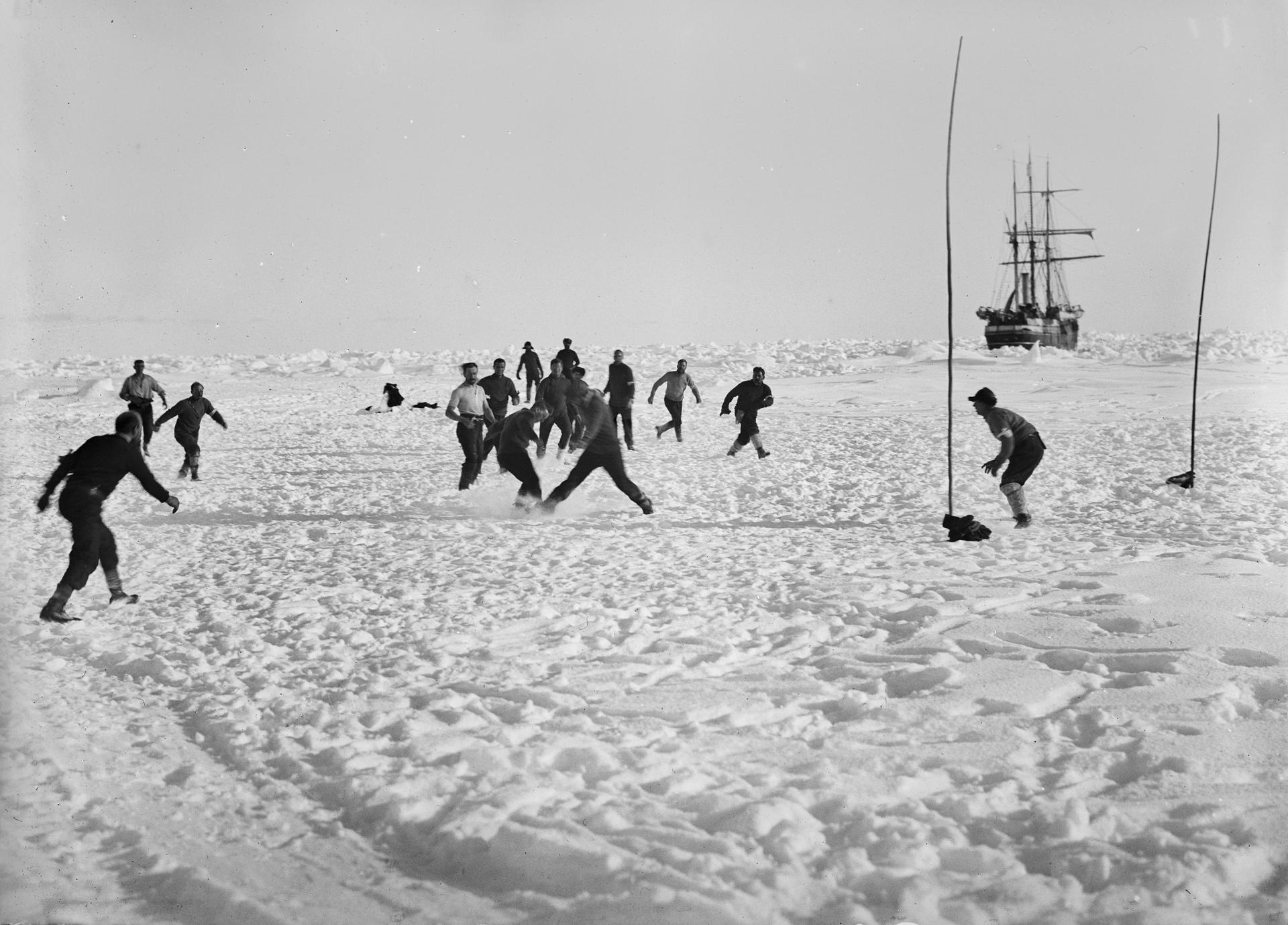 Men play soccer in white, snowy field nearby stuck ship