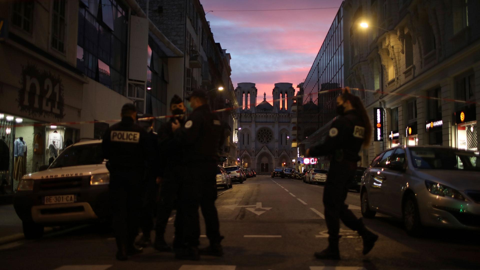 Several police are shown at the end of a street with the Notre Dame church in the distance and the sun setting.