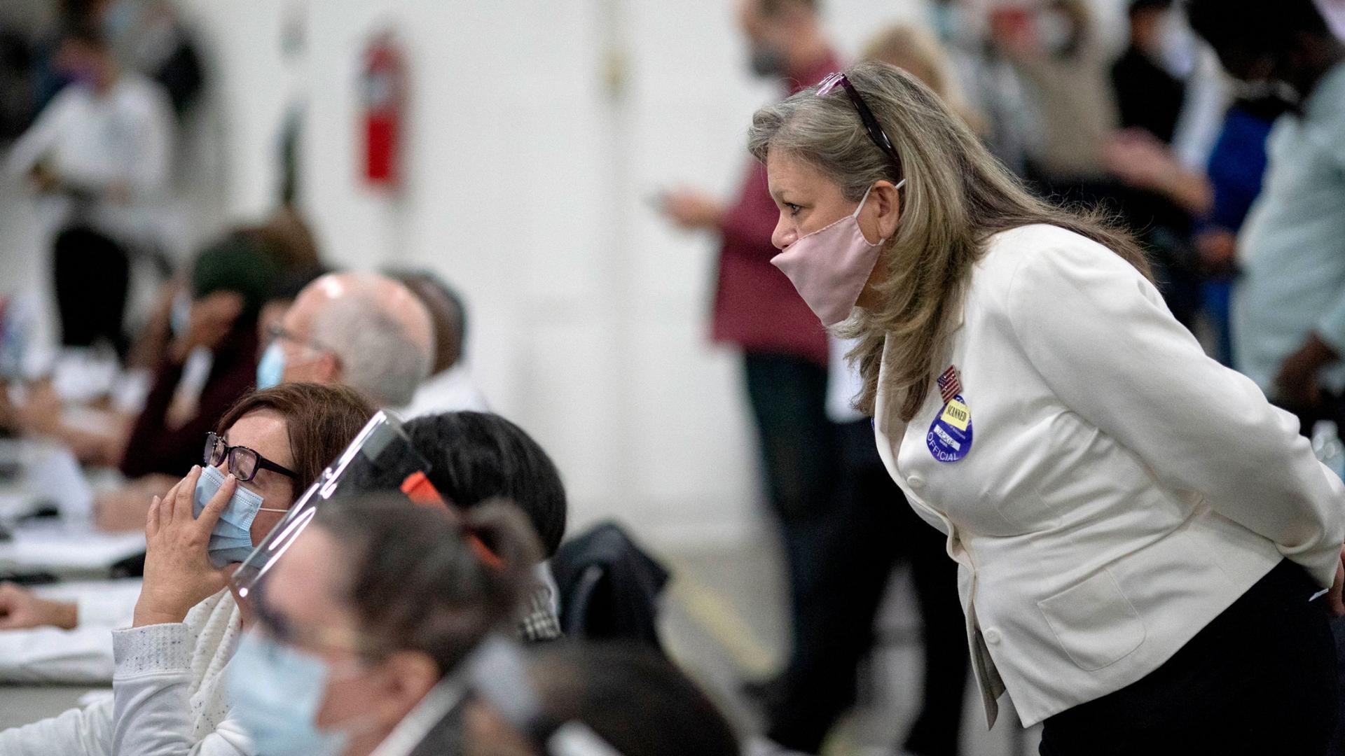 A woman wearing a white blazer and face mask is shown looking over the shoulder of a row of seated election inspectors.