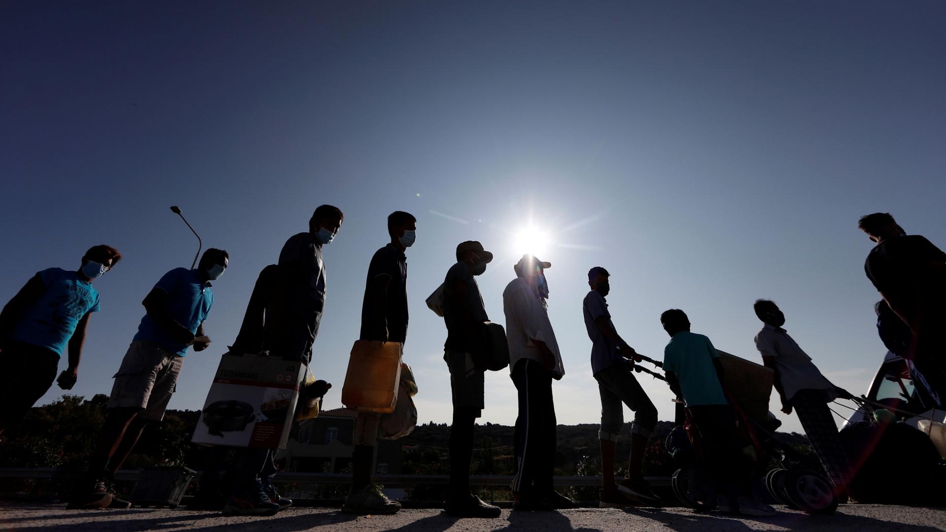 Refugees and migrants are shown in shadow standing in a lin carrying various belongings with the sun flaring in the distance.