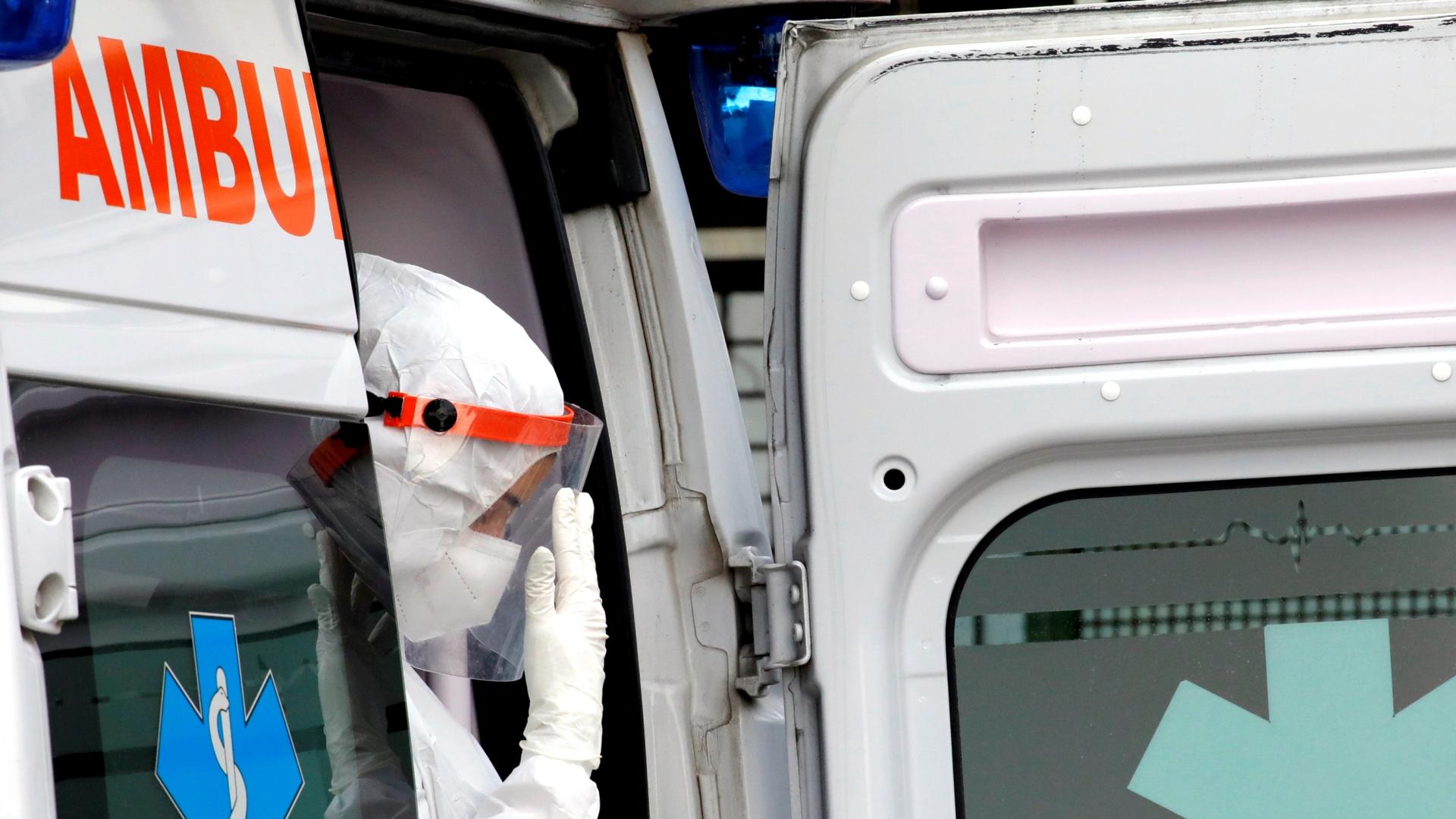 A person wearing white protective gear and gloves sits in the doorway of an ambulance. 