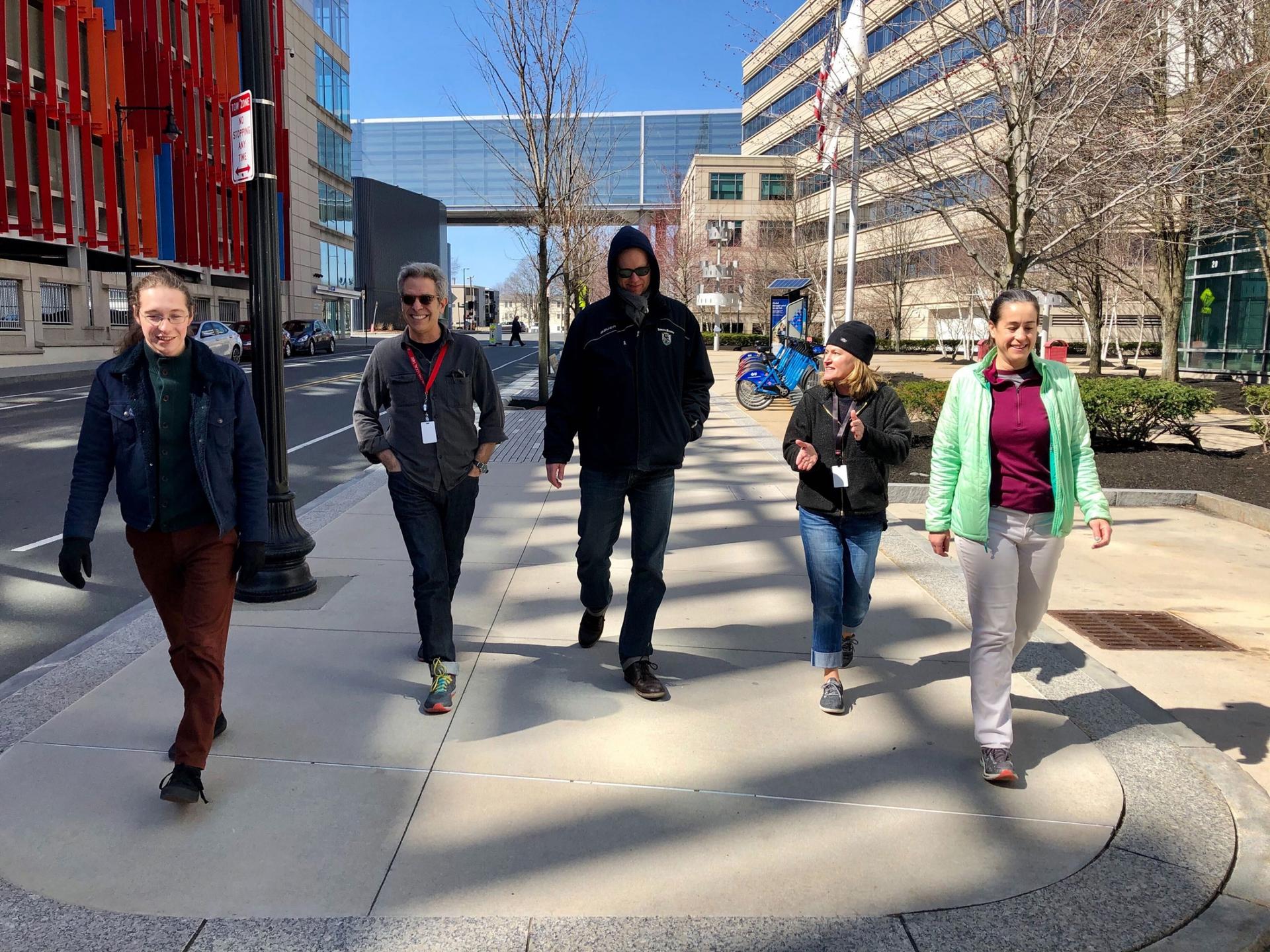 Five staff members from The World are shown walking on the sidewalk next to a colorful parking garage and the GBH headquarters building, west.
