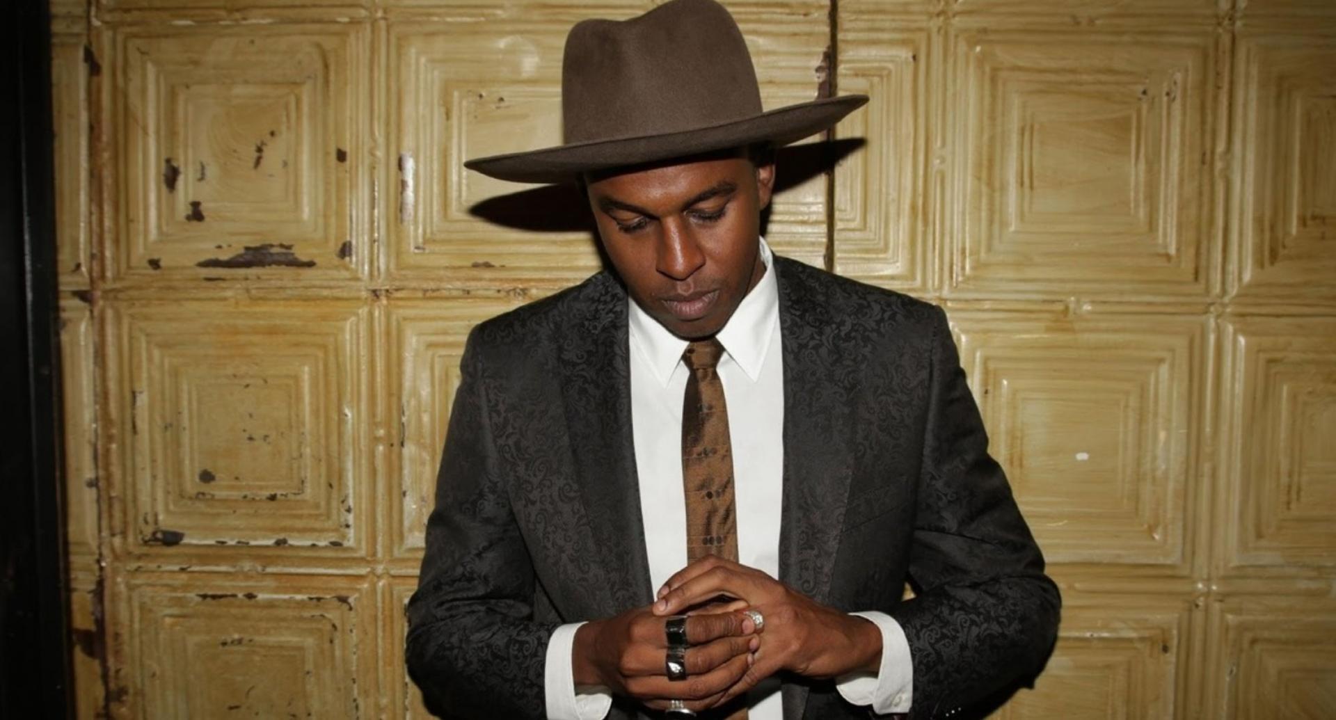 Sinkane is shown wearing a suit and wide-brimmed hat.