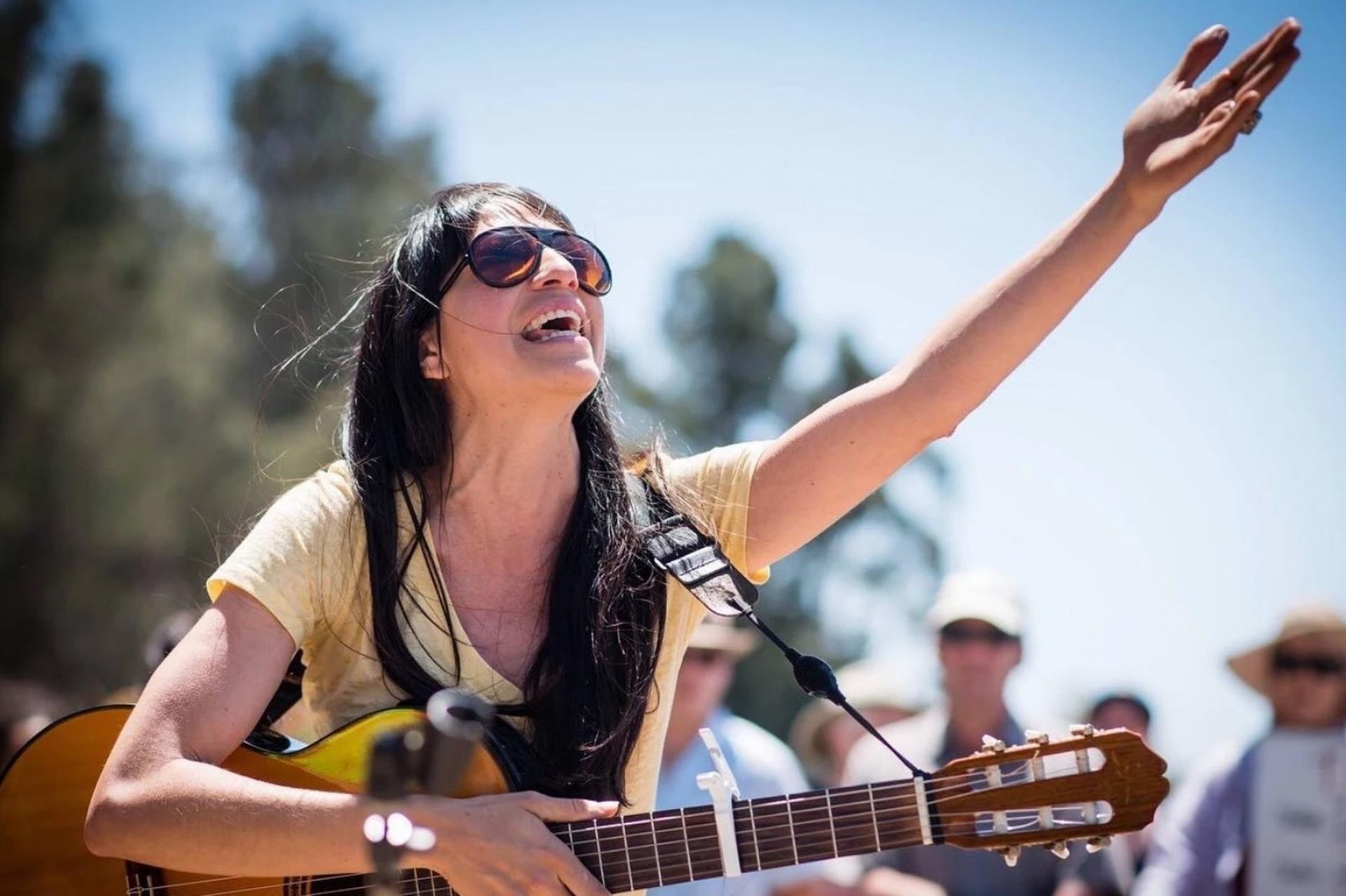 A woman wearing sunglasses holds a guitar in one hand while gesturing to the open blue sky with the other hand. 