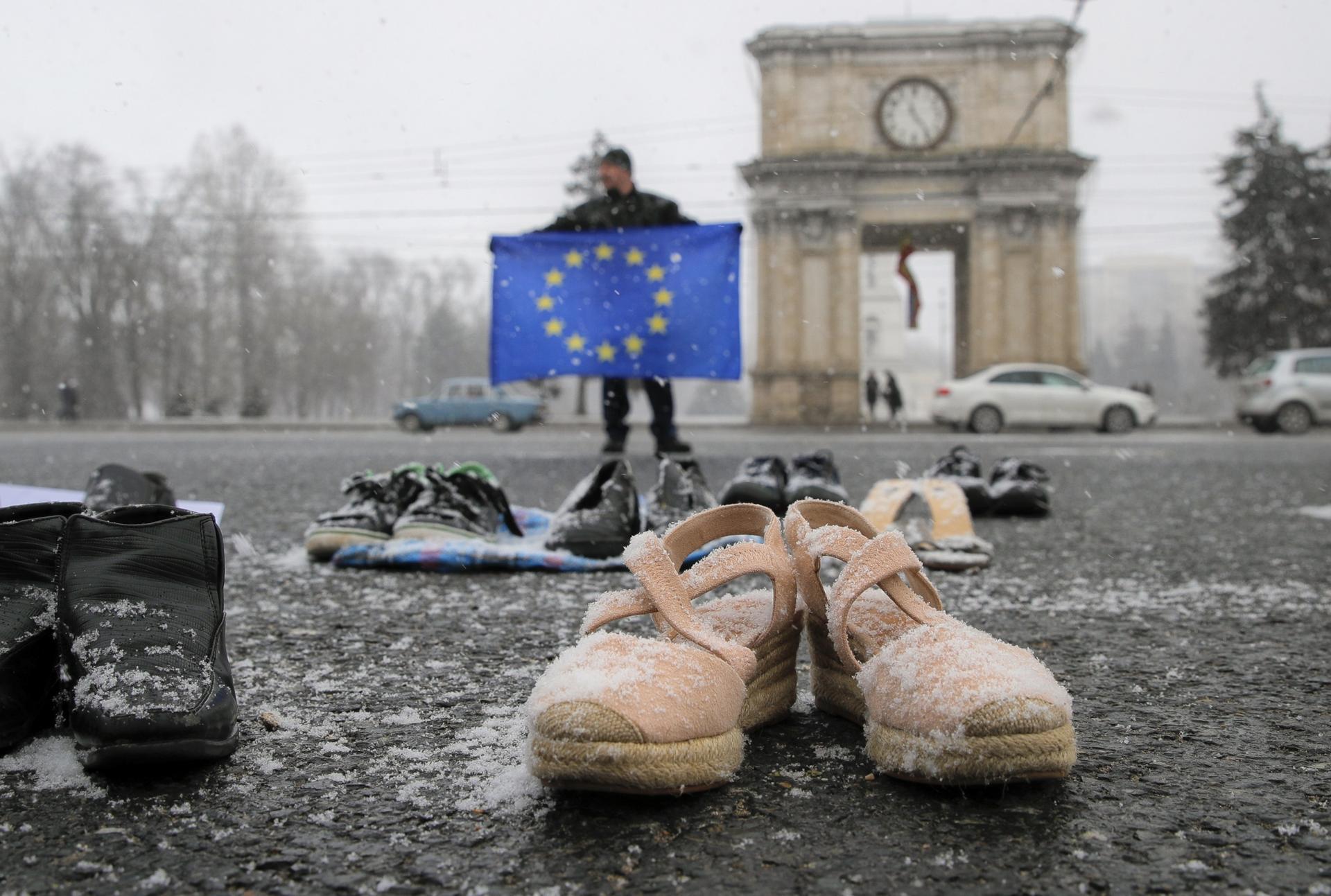 A few pairs of shoes with snow on them lie in a square near a man with an EU flag with blue and yellow colors. 