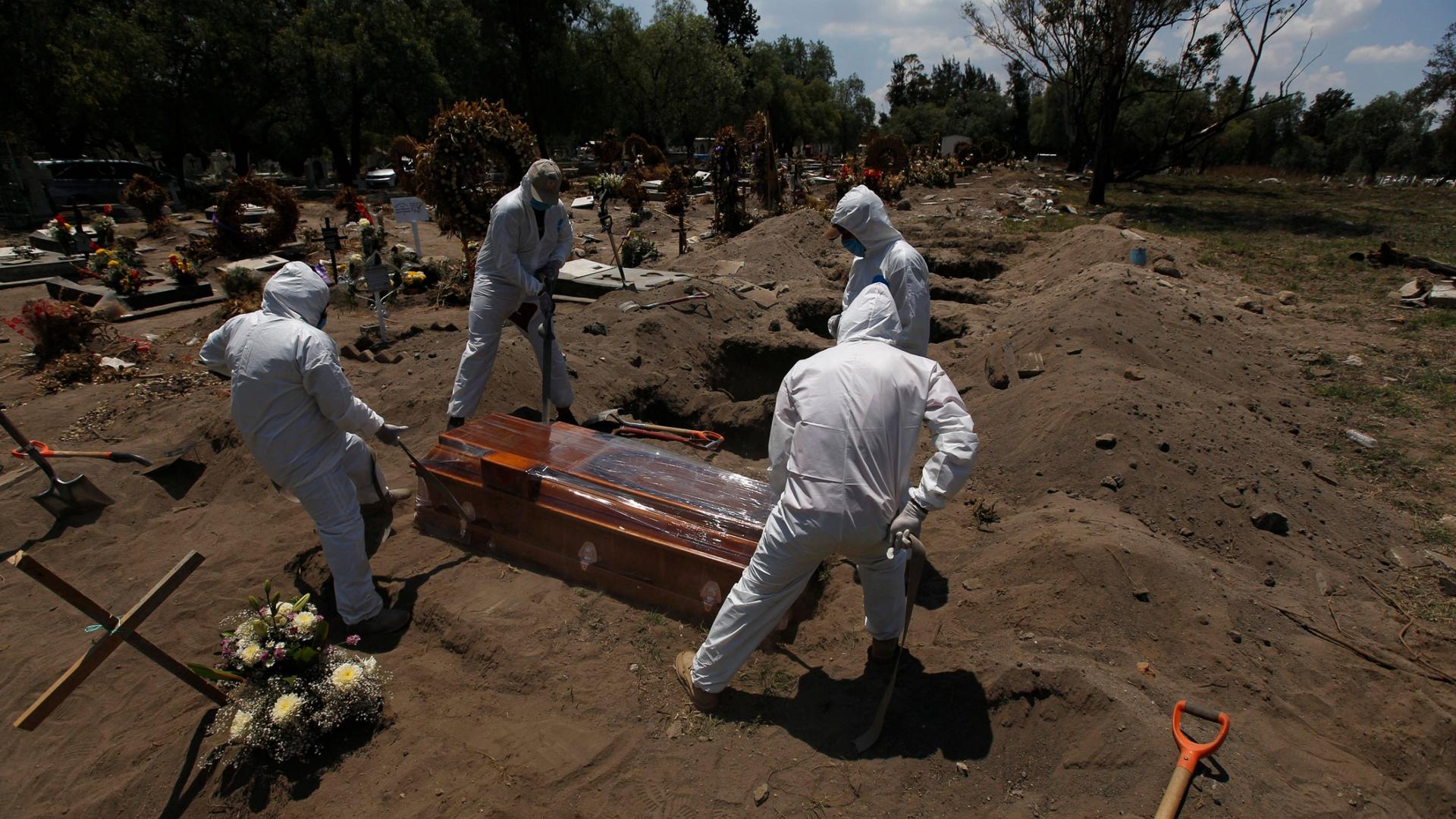 Four people are shown on holding ropes on each corner of a brown wooden coffin in a row with several dug out graves awaiting future burials.