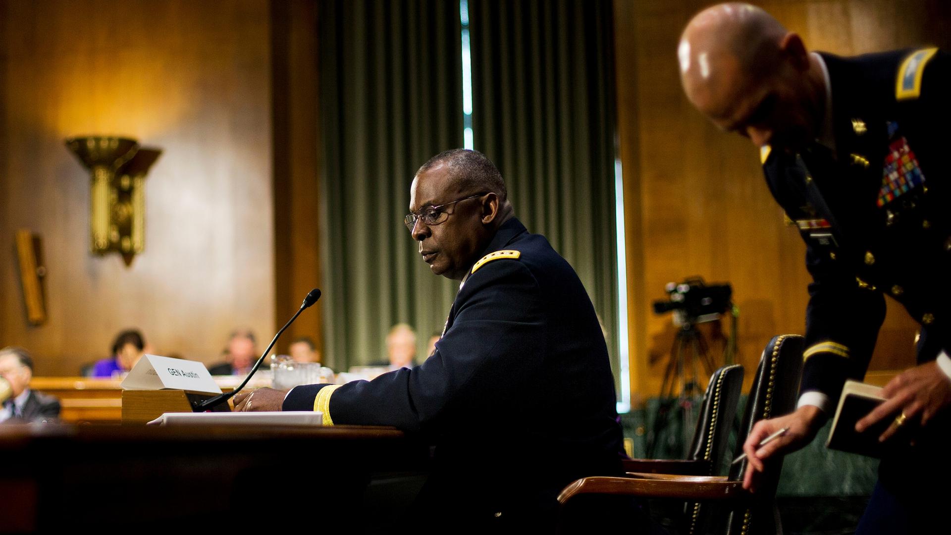 Then US Central Command Commander Gen. Lloyd Austin is shown seated at a large wooden table and behind a microphone.