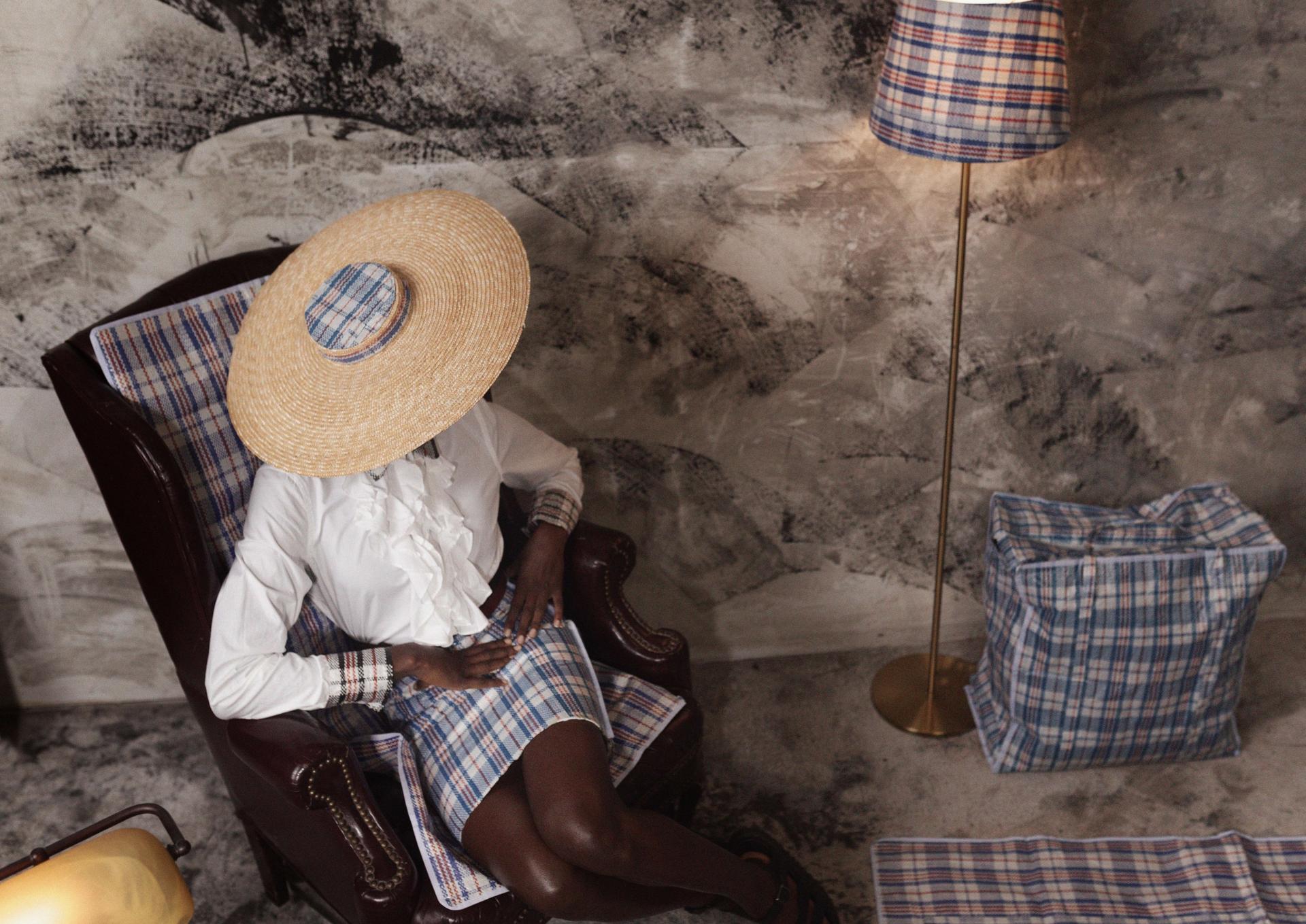 A woman sits wearing a hat, white blouse and plaid plastic skirt with lamp shade of same fabric near plaid travel bag