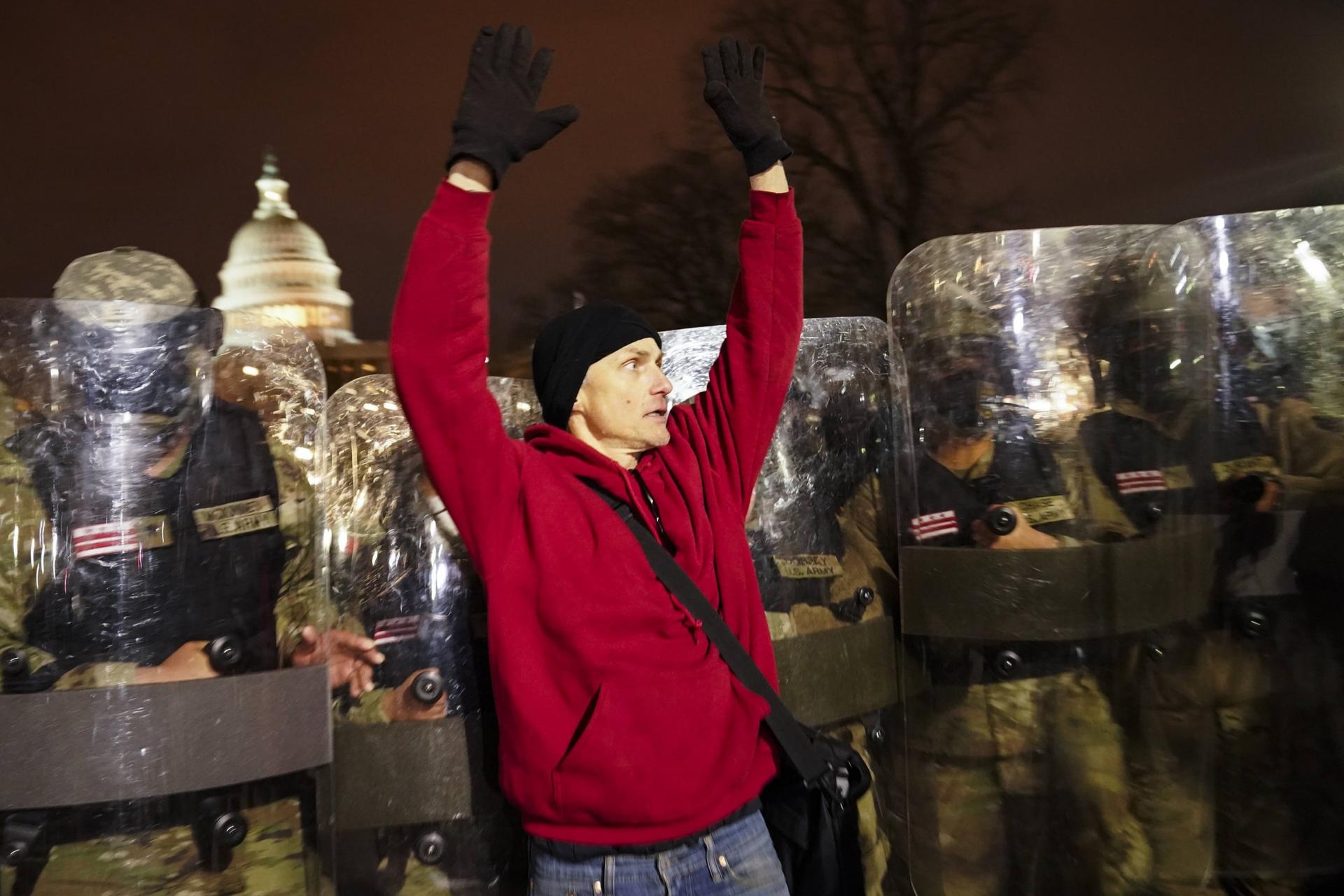 National guard appear behind a man wearing a red sweater, who has his hands up.