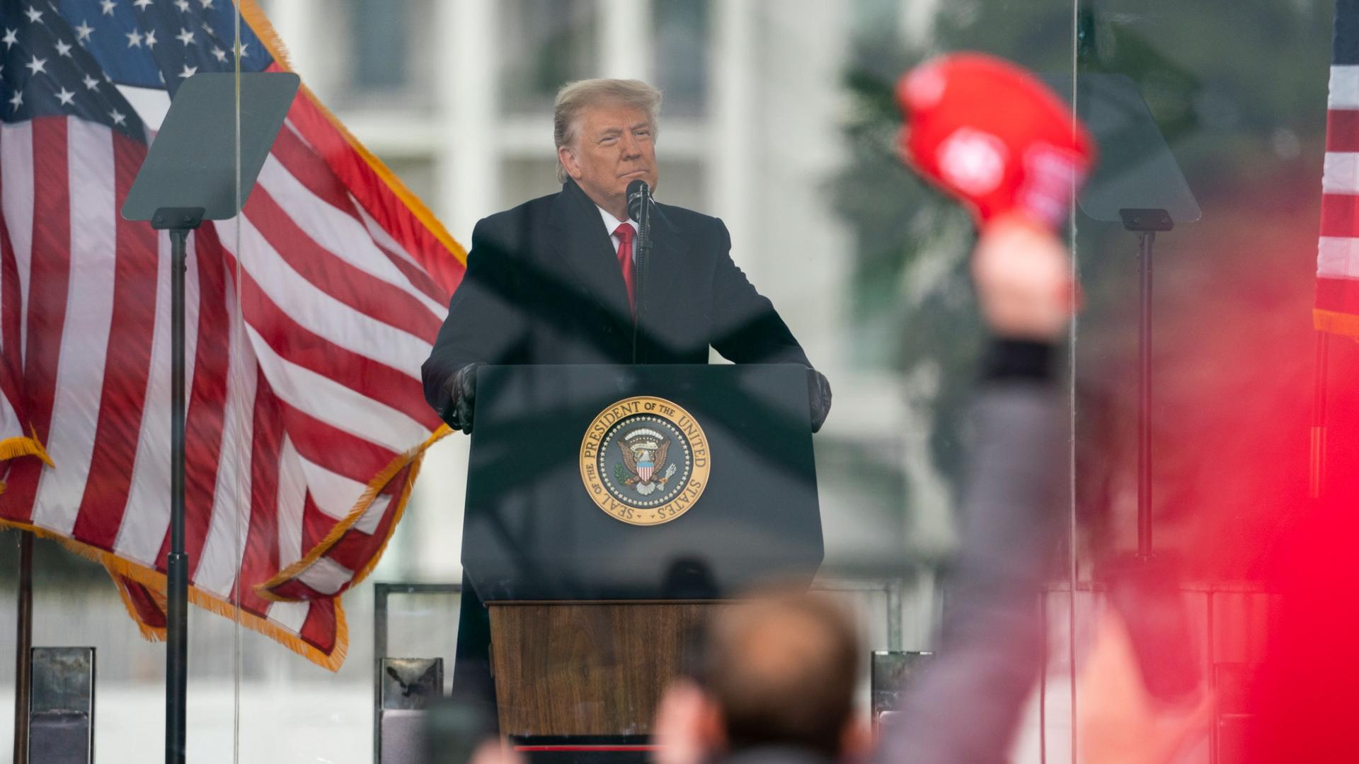 President Donald Trump is shown standing at a podium speaking with US flags waving behind him.