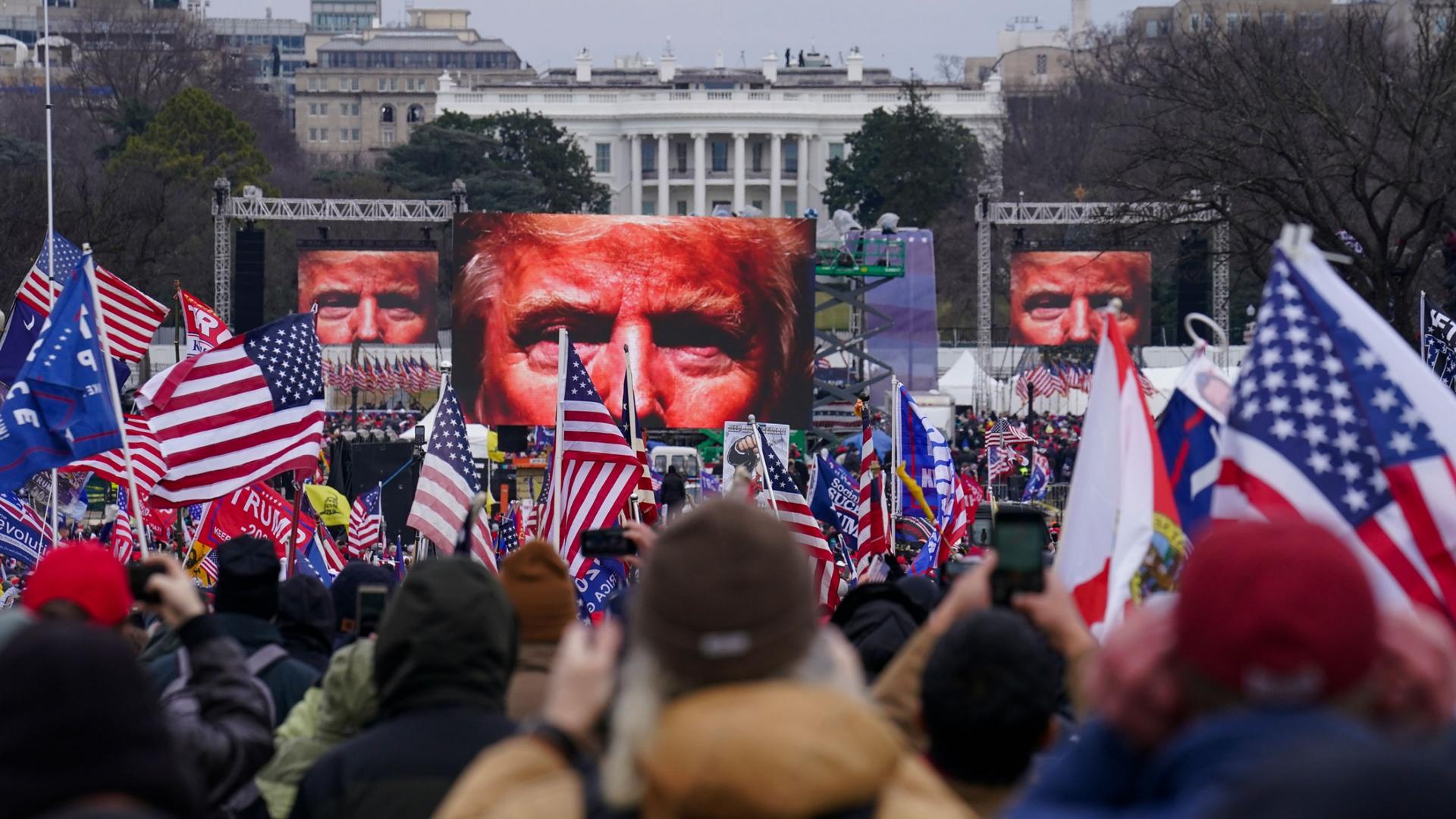 A large crowd of people are shown holding small US flags with screens showing Donald Trump's eyes in the distance.